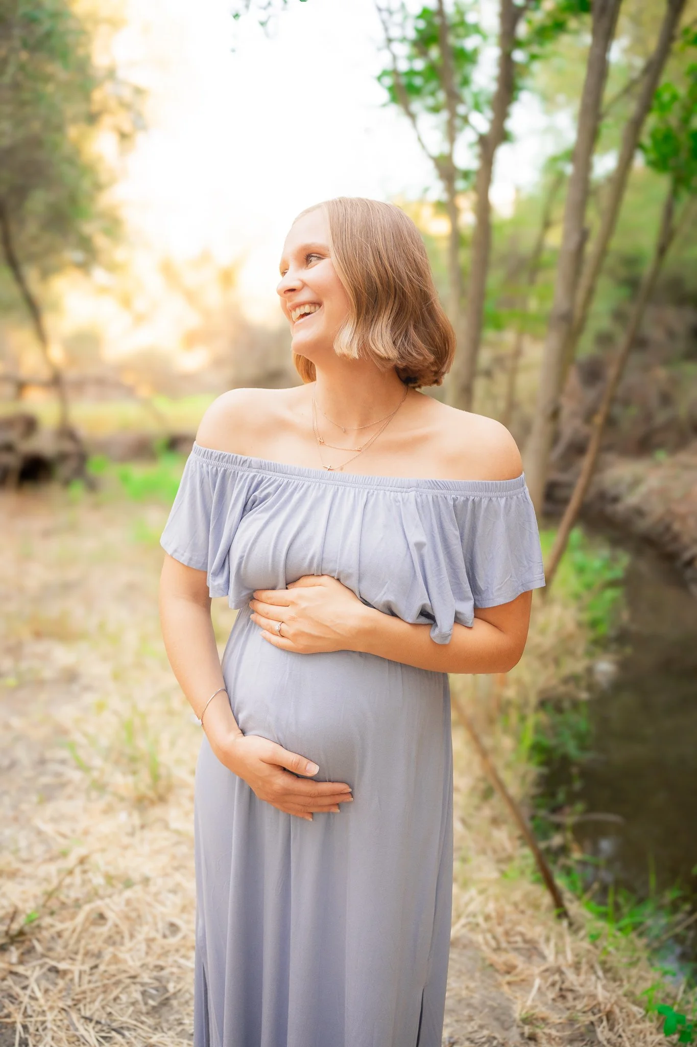 A pregnant woman in a gray off-the-shoulder dress stands outdoors, smiling and looking to her left, with trees and sunlight in the background.