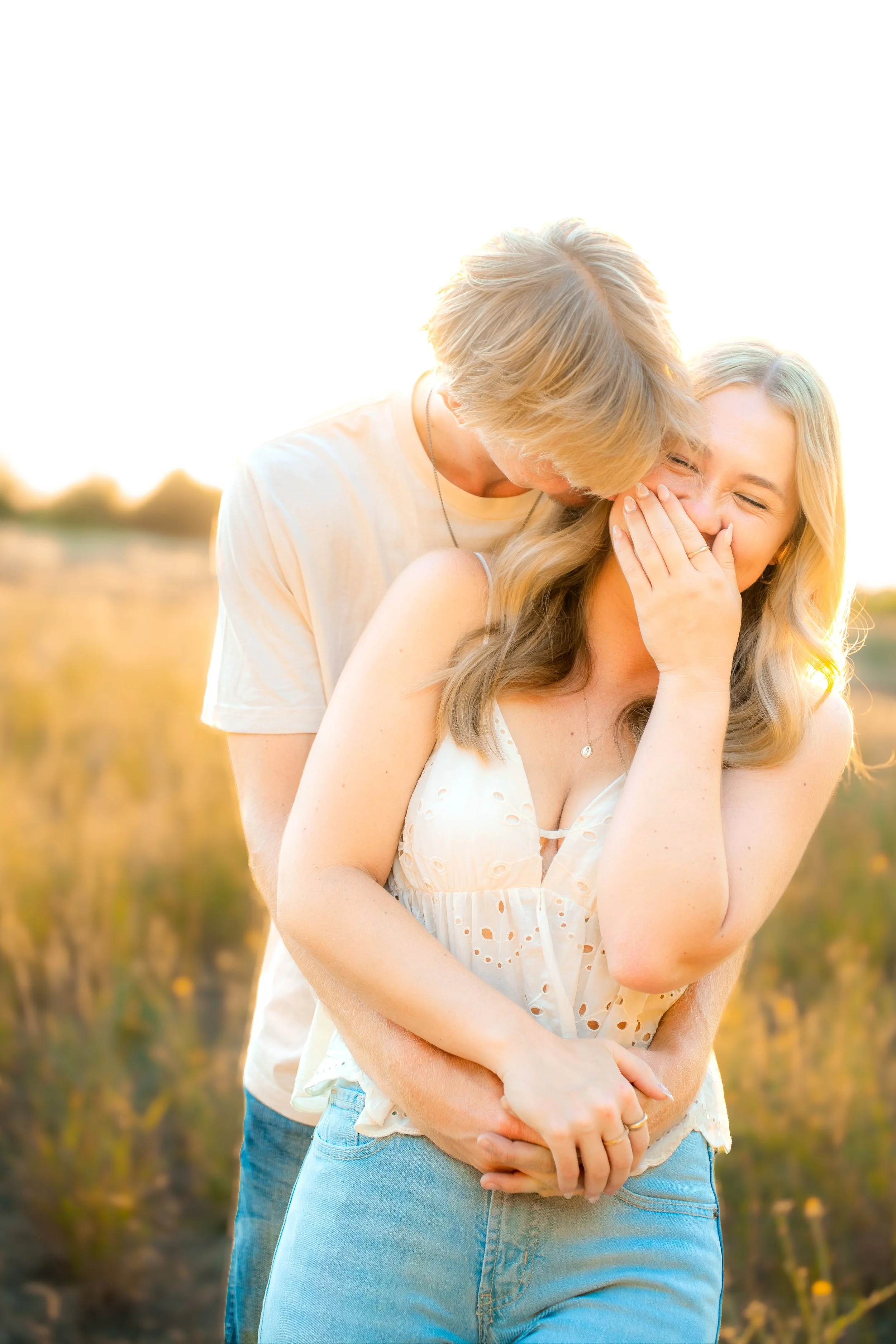A young couple outdoors in a field during sunset, with the man kissing the woman on the cheek as she laughs, both wearing light-colored clothing during a couples session in Bidwell Park in Chico, CA.