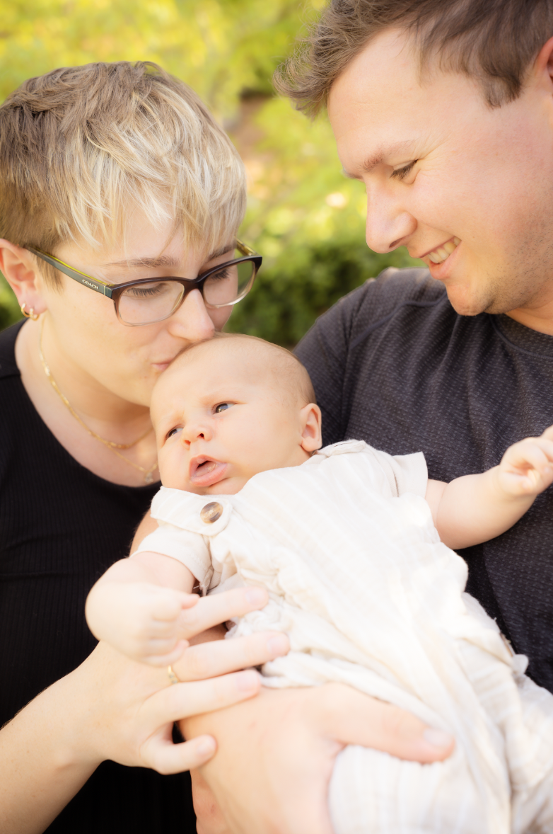 A woman and a man holding and smiling at a baby outdoors on a sunny day.