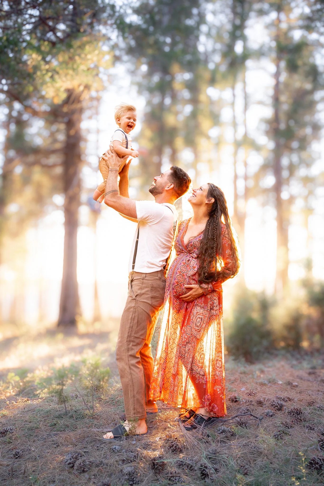 A happy family of three in a forest during sunset, with a man lifting a young boy and a woman smiling at each other.