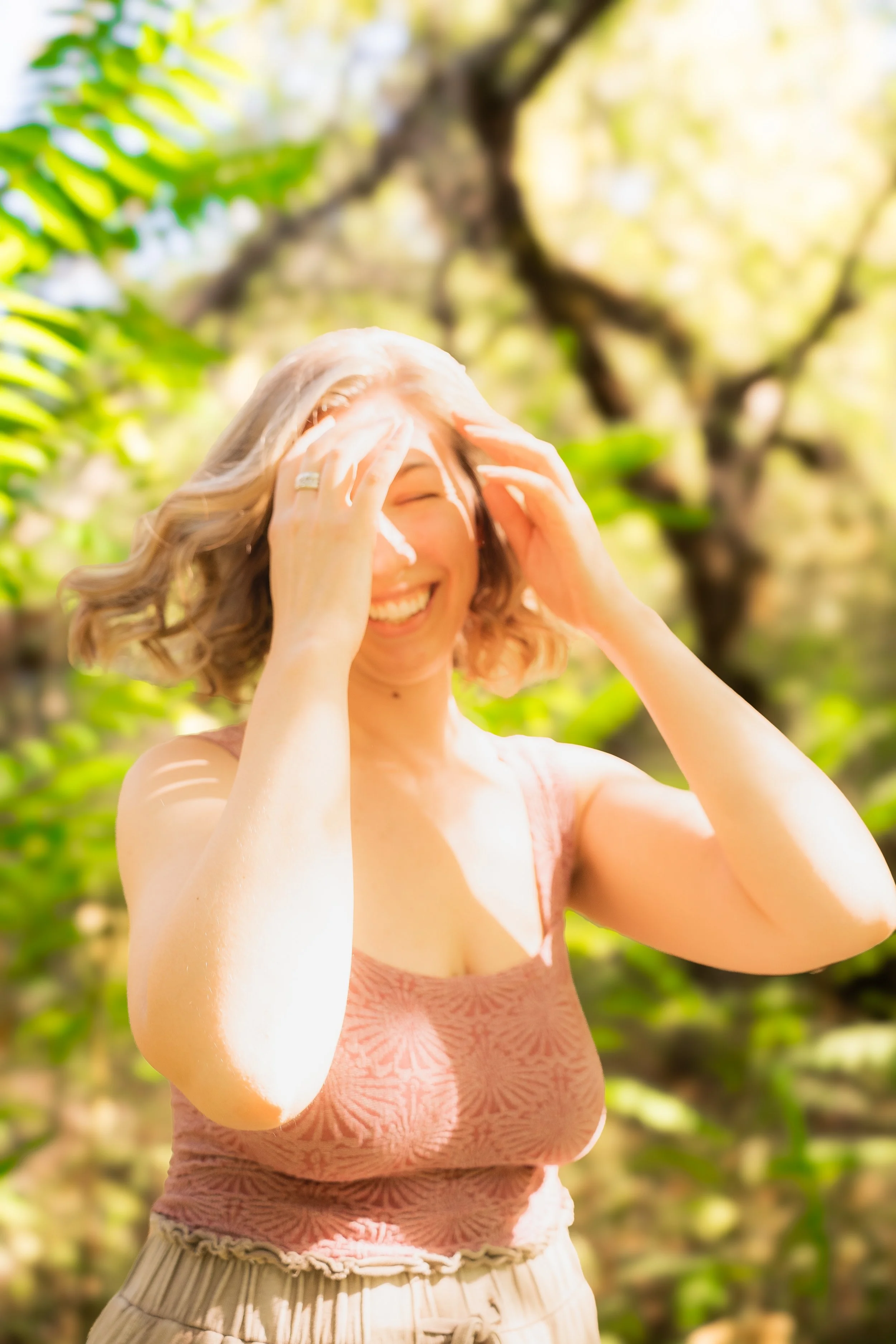 A young woman with blonde hair smiling and laughing outdoors, with trees and green foliage in the background.