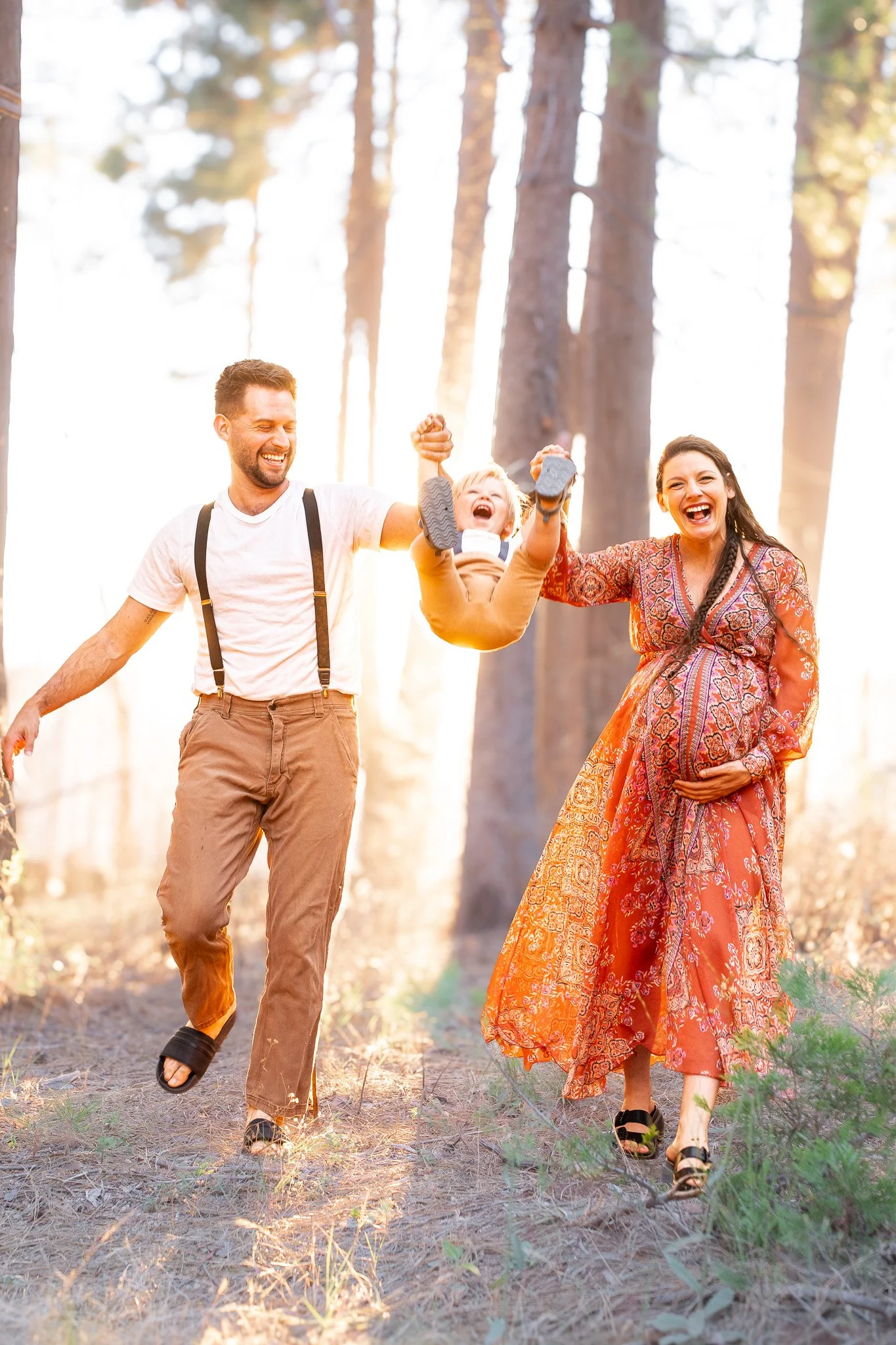 A family of three, a man, woman, and a young boy, enjoying a walk in a forest during the daytime. The boy is being lifted up by both parents. They are all smiling and laughing, dressed casually, with the woman wearing a colorful dress and the man in 