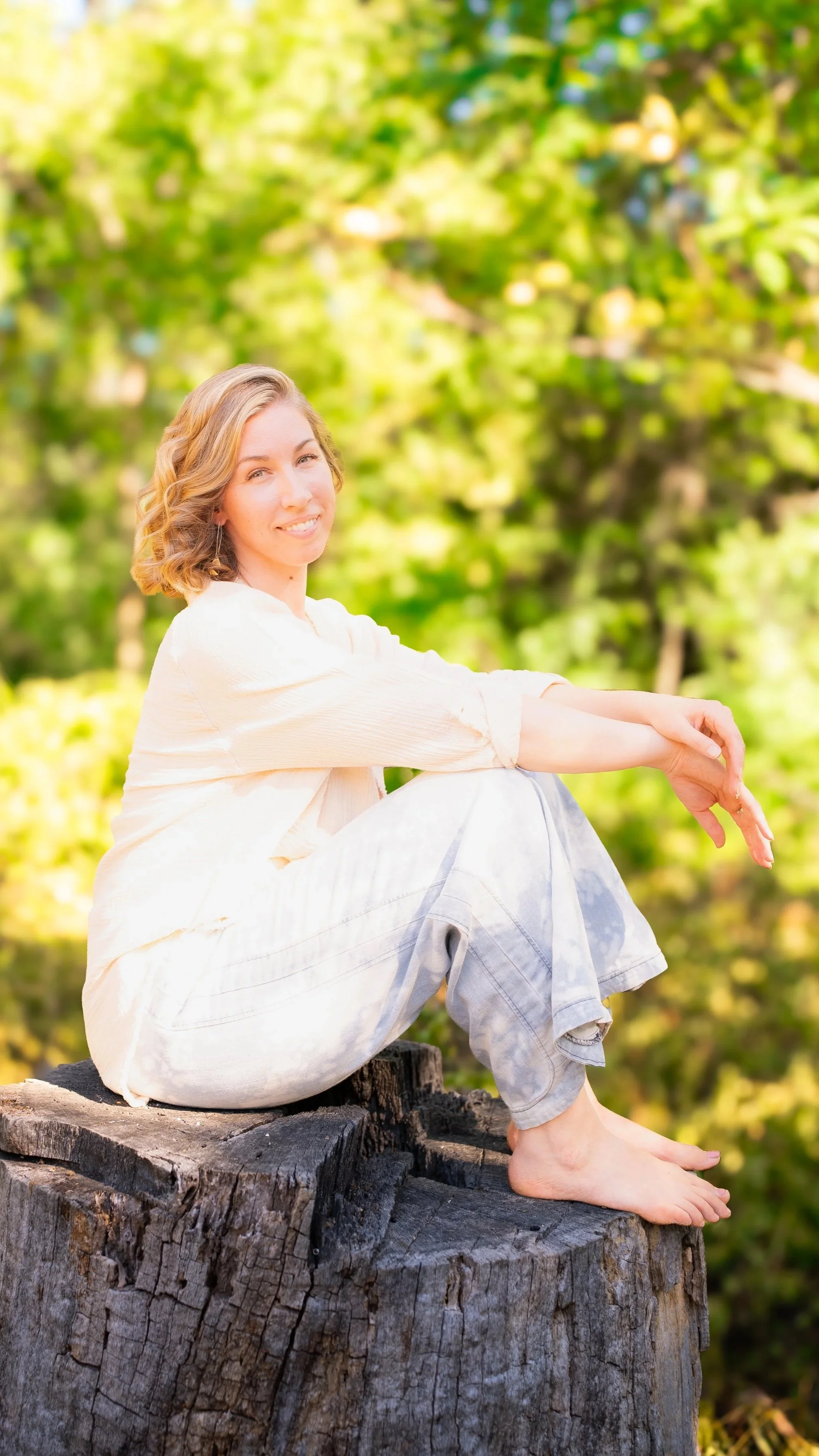 A woman with short curly hair sitting on a tree stump in a park with green trees in the background, smiling and looking at the camera.
