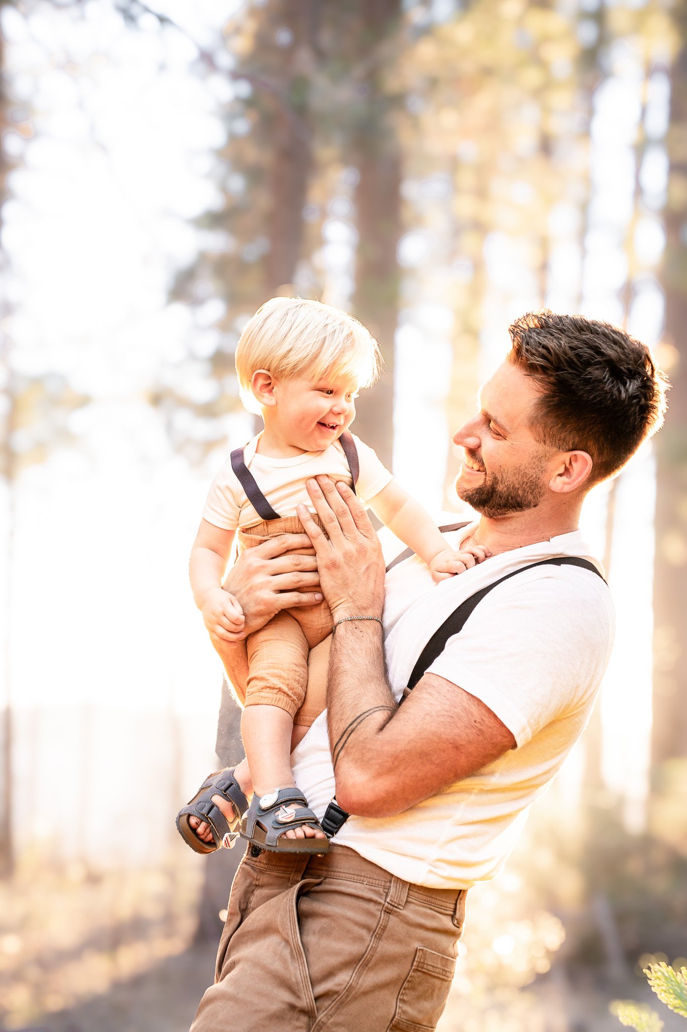 A man with short dark hair and a beard smiling and holding a young boy with blonde hair, dressed in a beige t-shirt, dark sandals, and brown pants, outdoors in a sunny, wooded area.