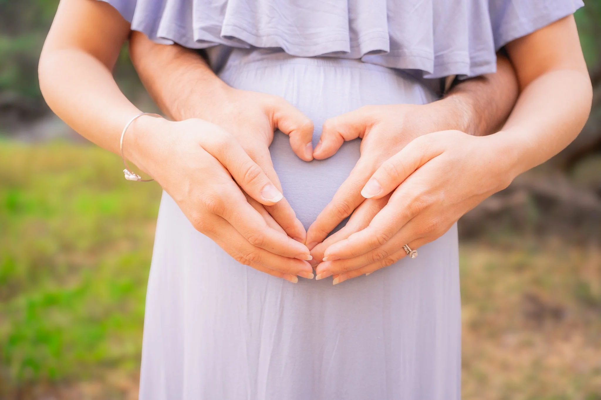 Close-up of a pregnant woman with a man's hands forming a heart shape on her belly, outdoors with blurred greenery in the background.