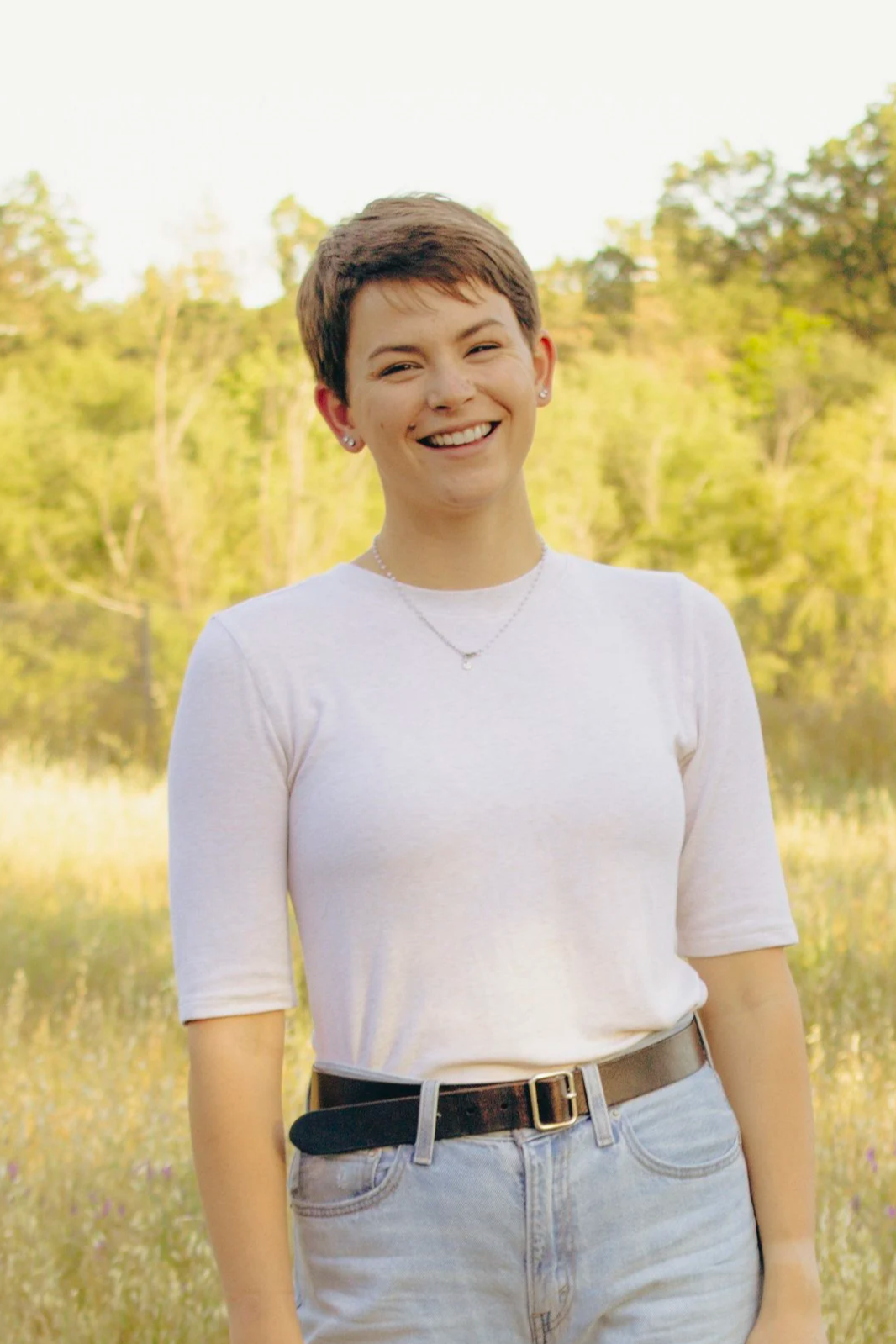 A smiling young woman with short brown hair, wearing a white t-shirt and light-colored jeans, standing outdoors near a body of water with trees and greenery in the background.