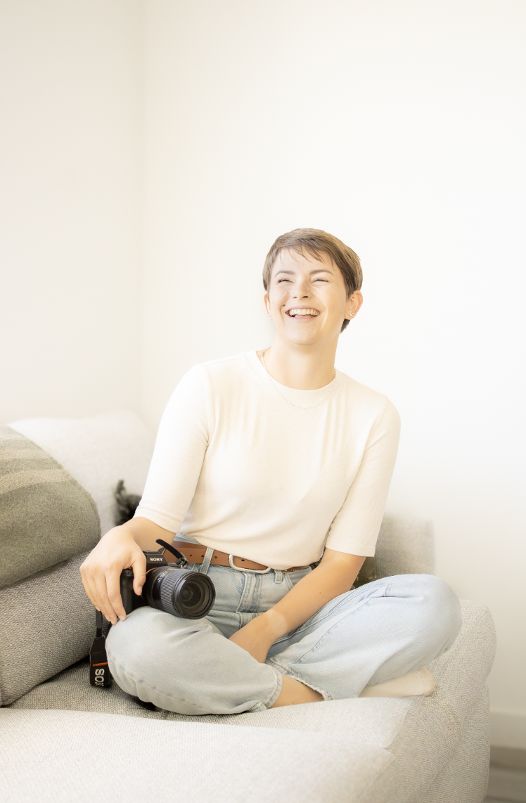 A woman sitting on a couch laughing and holding a camera in her hand.
