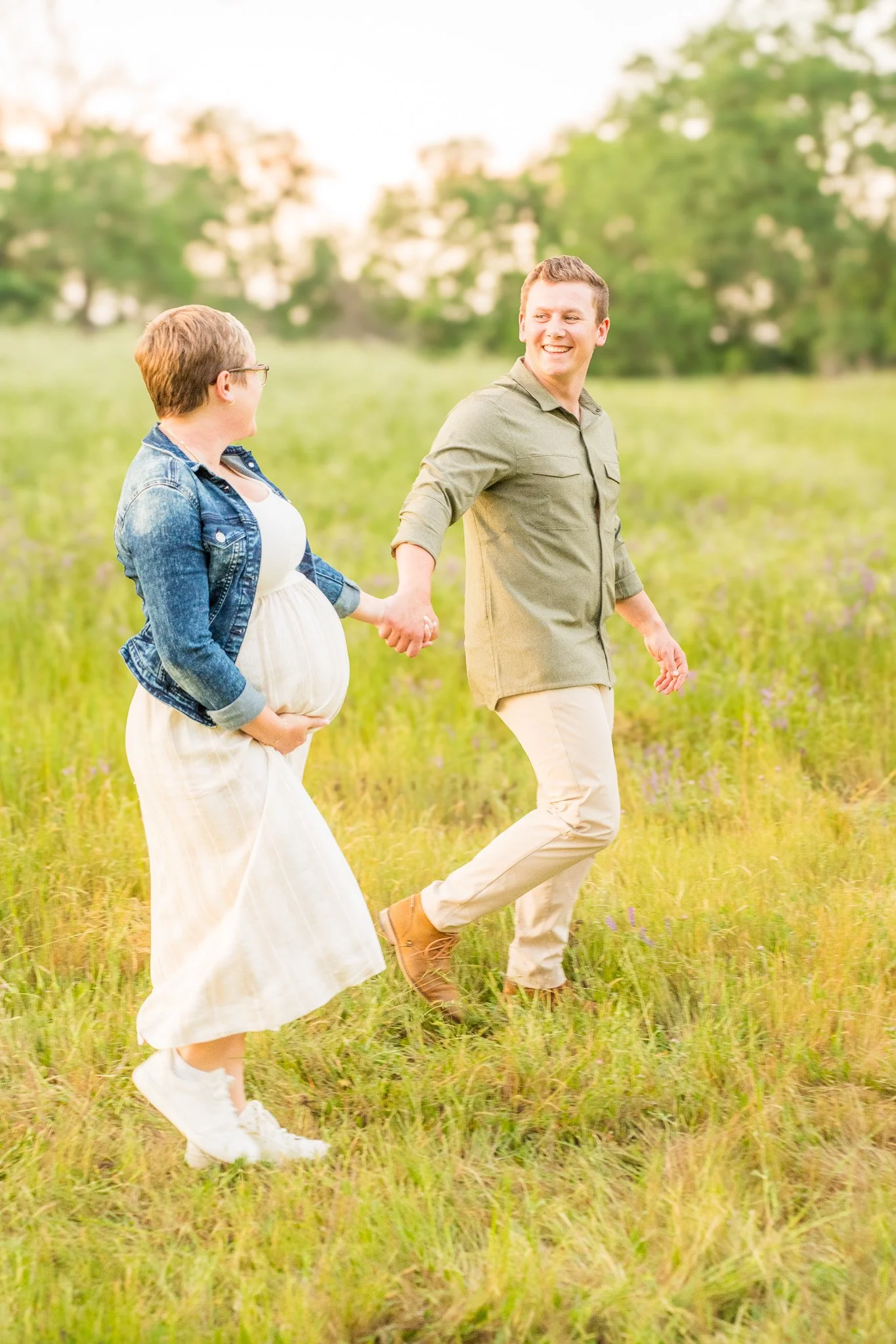 A couple holding hands and walking through a grassy field on a sunny day, with trees in the background.