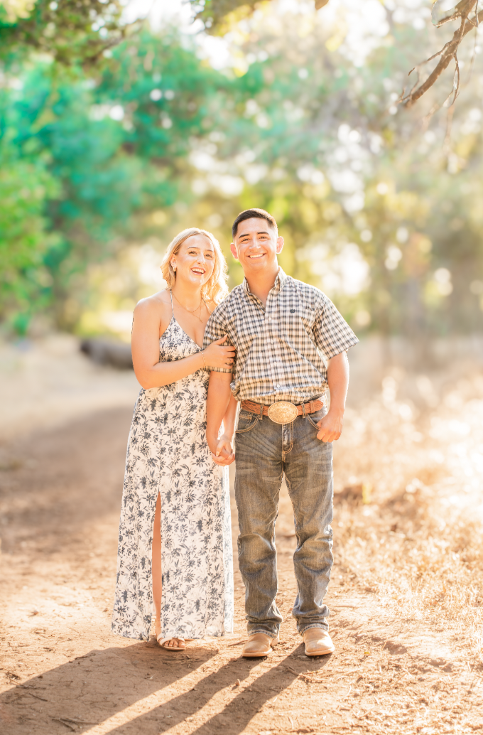 A smiling couple walking hand in hand outdoors on a dirt path, surrounded by trees with sunlight filtering through the leaves.