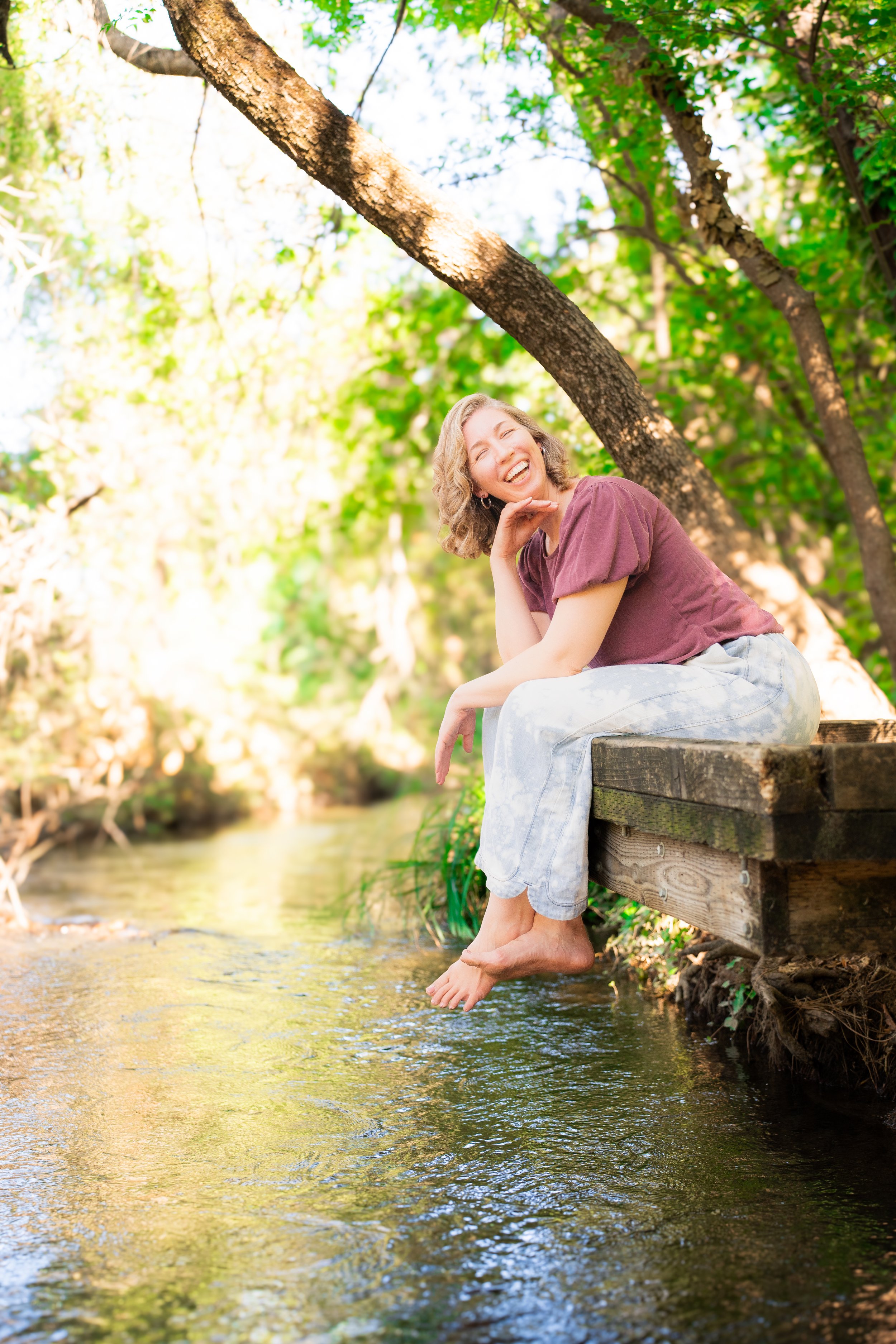 A woman sitting on a wooden dock with her feet dangling in a small creek, smiling and enjoying the sunny day surrounded by green trees.
