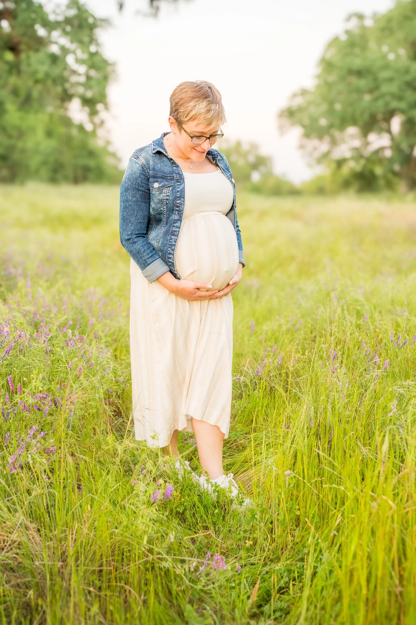 Pregnant woman standing in a green field with purple flowers, wearing a denim jacket and a cream-colored dress, smiling and looking down.