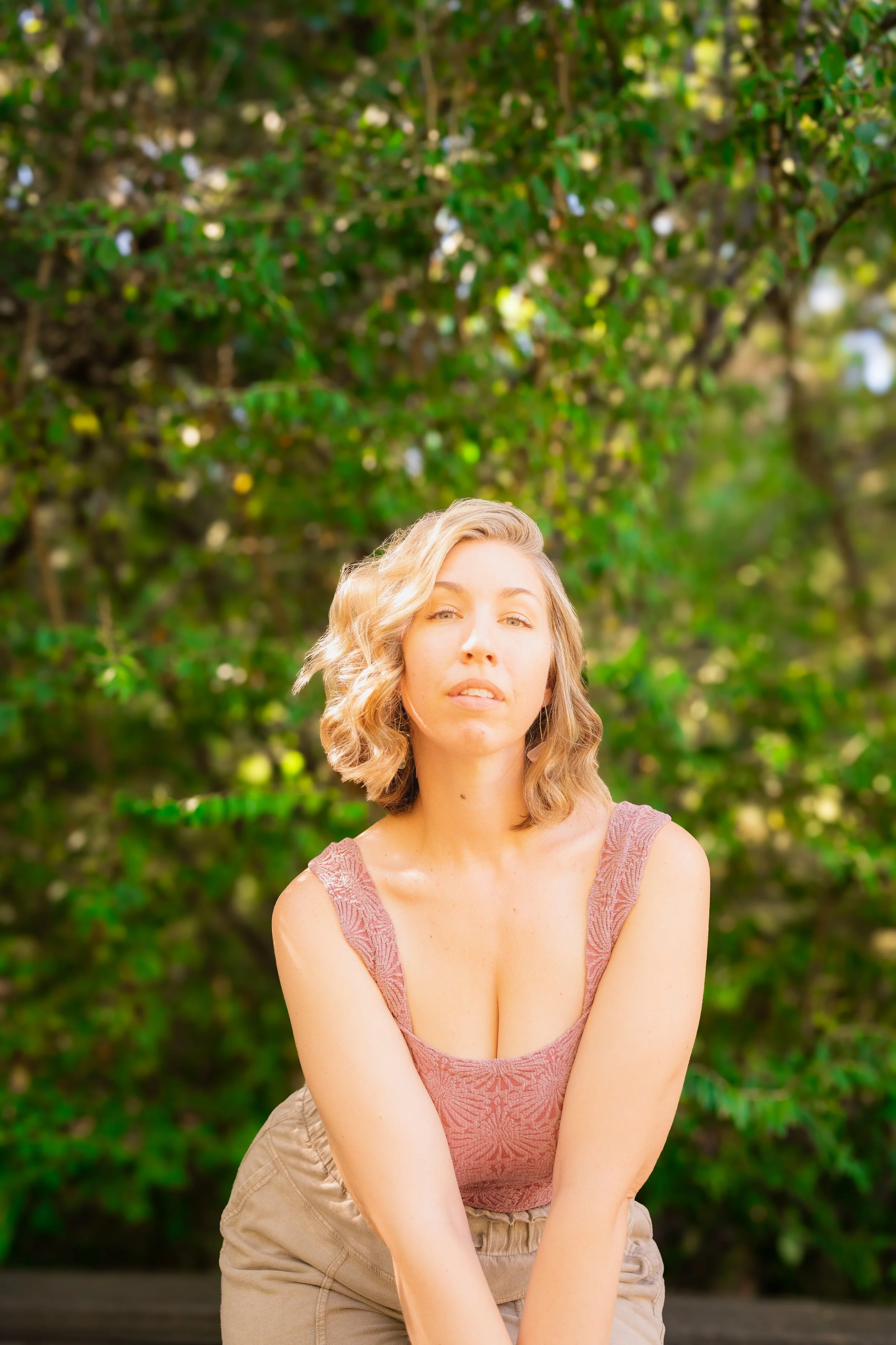 A woman with wavy blonde hair wearing a pink lace tank top and khaki pants, standing outdoors in front of a leafy, green background.