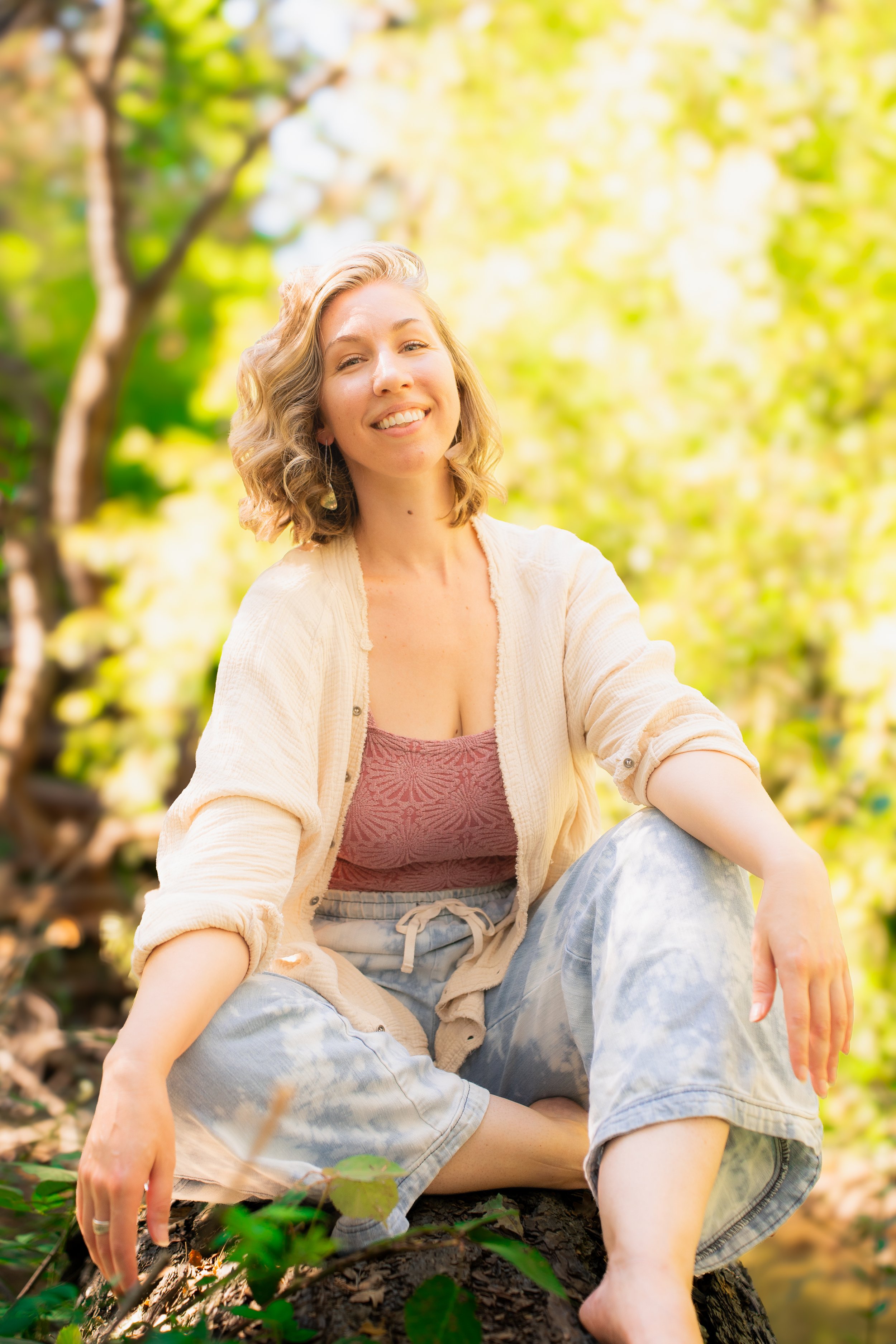 A woman with curly blonde hair smiling and sitting on a fallen tree in a lush green forest during daytime.