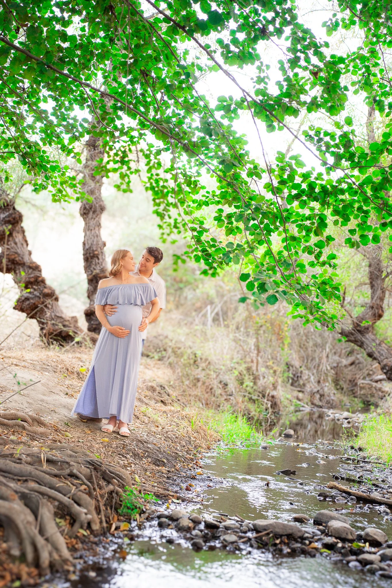 A pregnant woman in a light gray dress, standing by a stream in a wooded area, with a man behind her, both looking at each other lovingly.