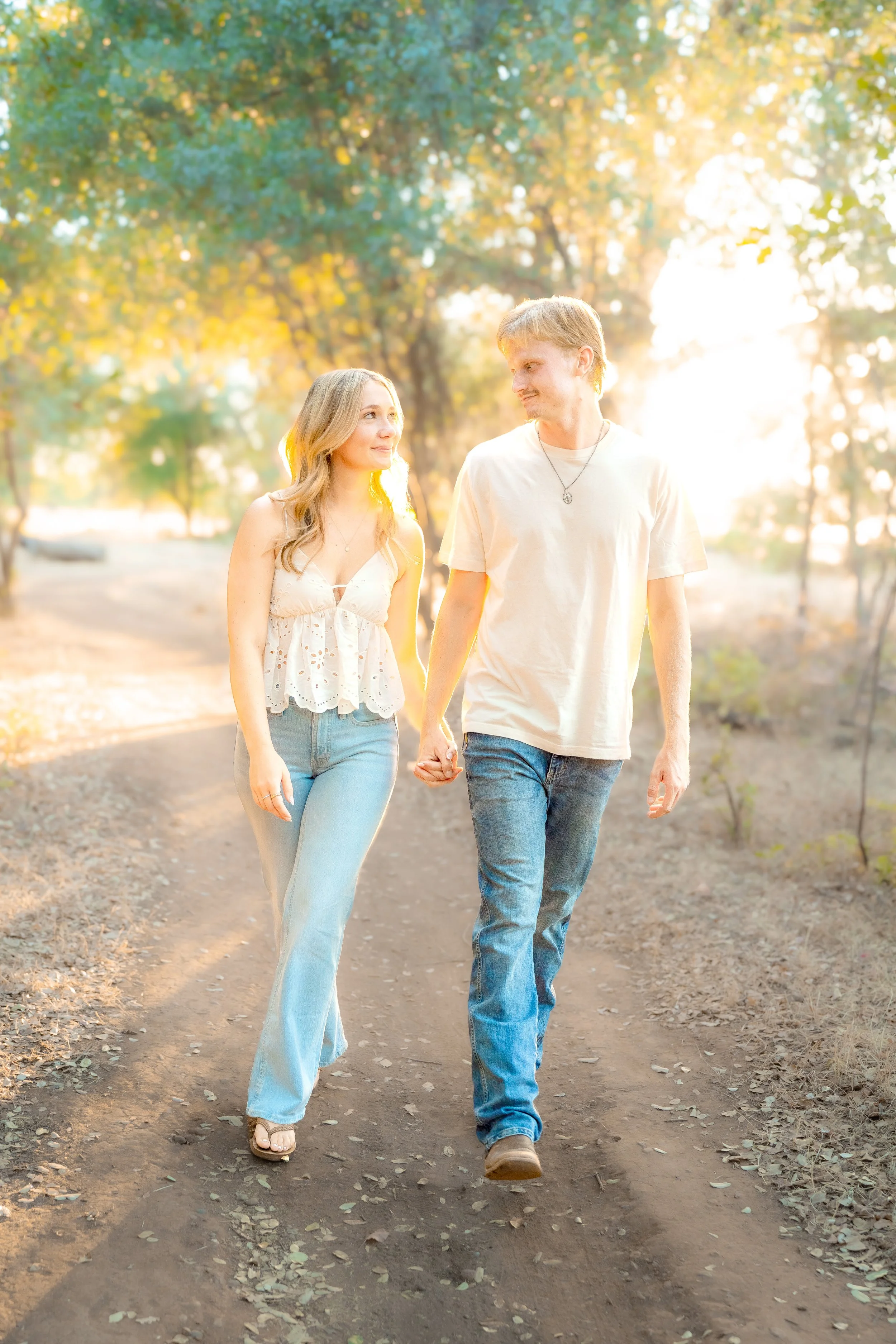 A young couple holding hands and walking together outdoors on a dirt path during sunset, surrounded by trees with green and yellow leaves during a couples session in Bidwell Park in Chico, CA.