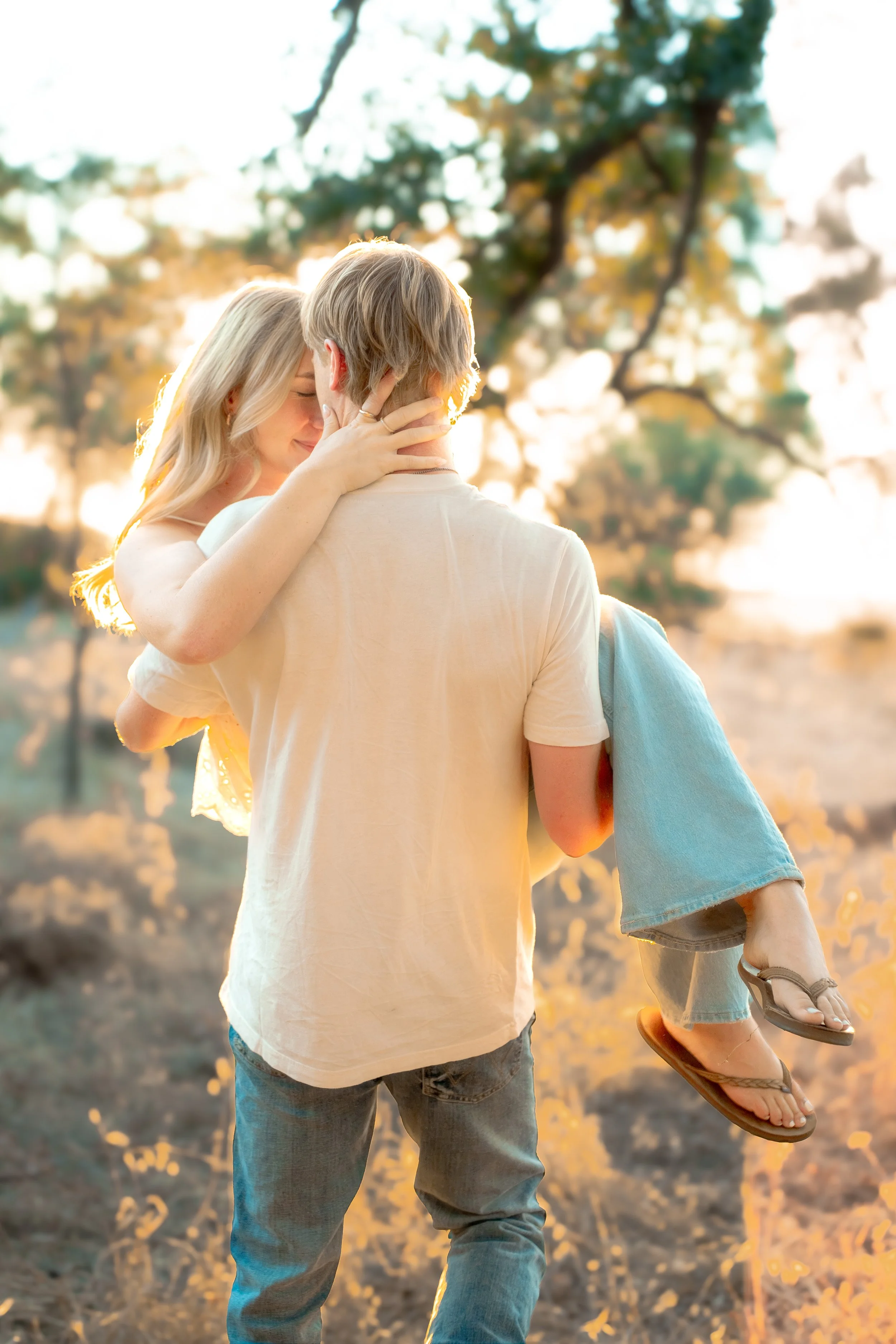 A couple embraces outdoors in warm sunlight, with the woman holding onto the man's neck as he carries her, both smiling with a background of trees and golden light during a couples session in Bidwell Park in Chico, CA.