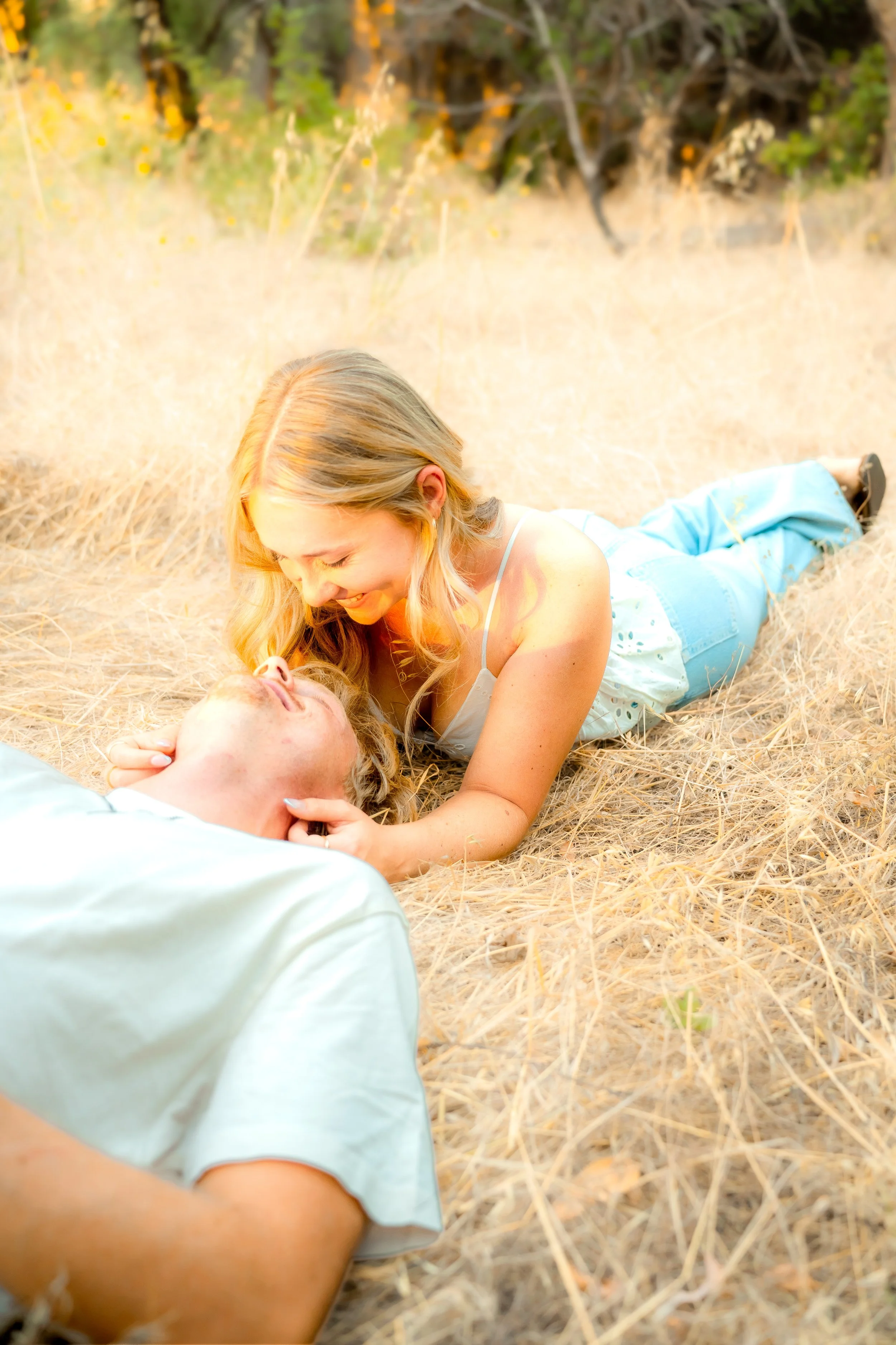 A woman with long blonde hair, wearing a white tank top and blue jeans, lying on her stomach in a field of dry grass, smiling and leaning over a person laying on the ground during a couples session in Bidwell Park in Chico, CA.