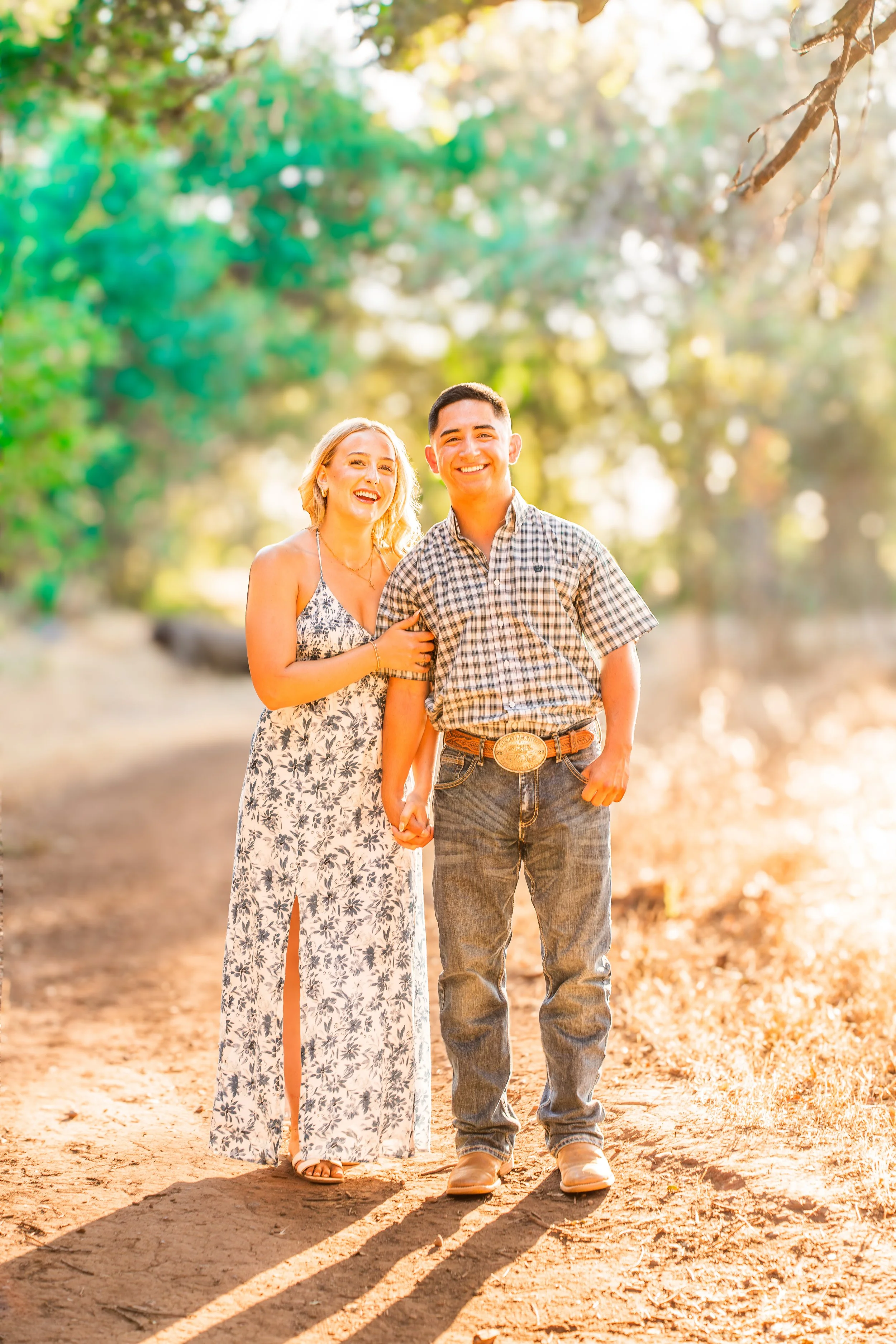 A smiling couple walking hand-in-hand on a dirt path in a wooded area during sunset, both dressed in casual summer clothing during a couples session in Bidwell Park in Chico, CA.