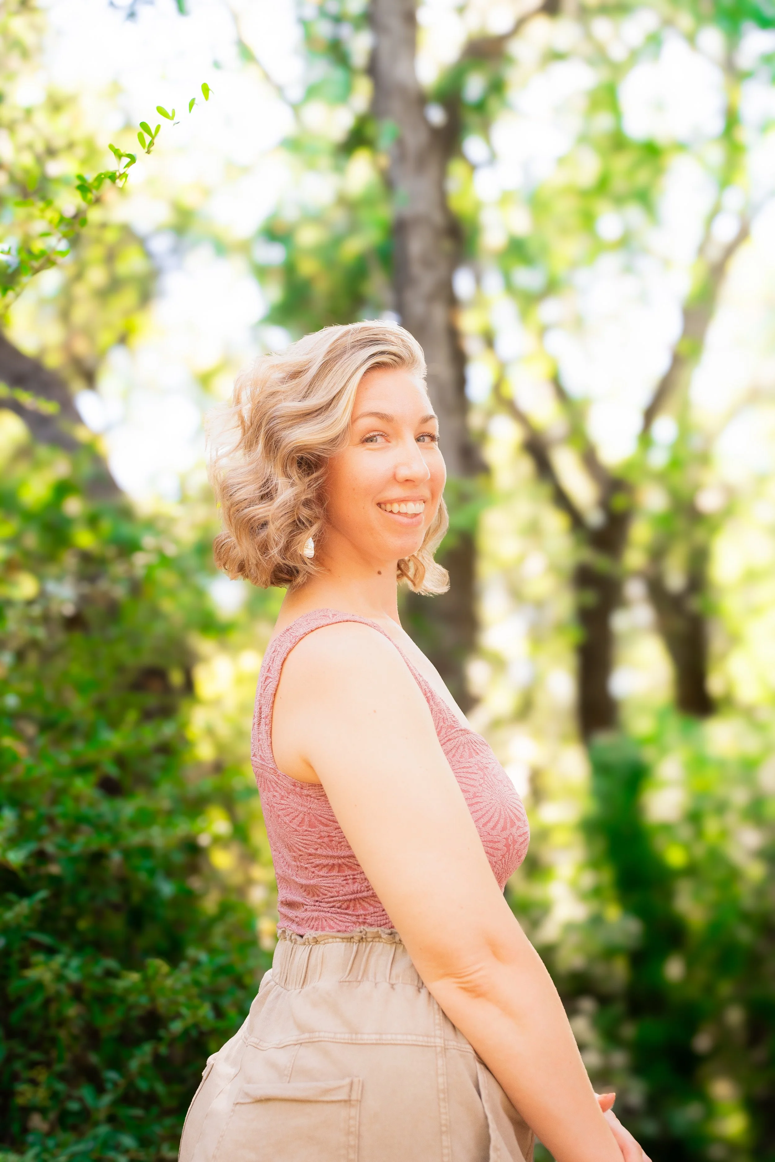 A woman with curly blonde hair smiling outdoors in a park-like setting with green trees in the background.