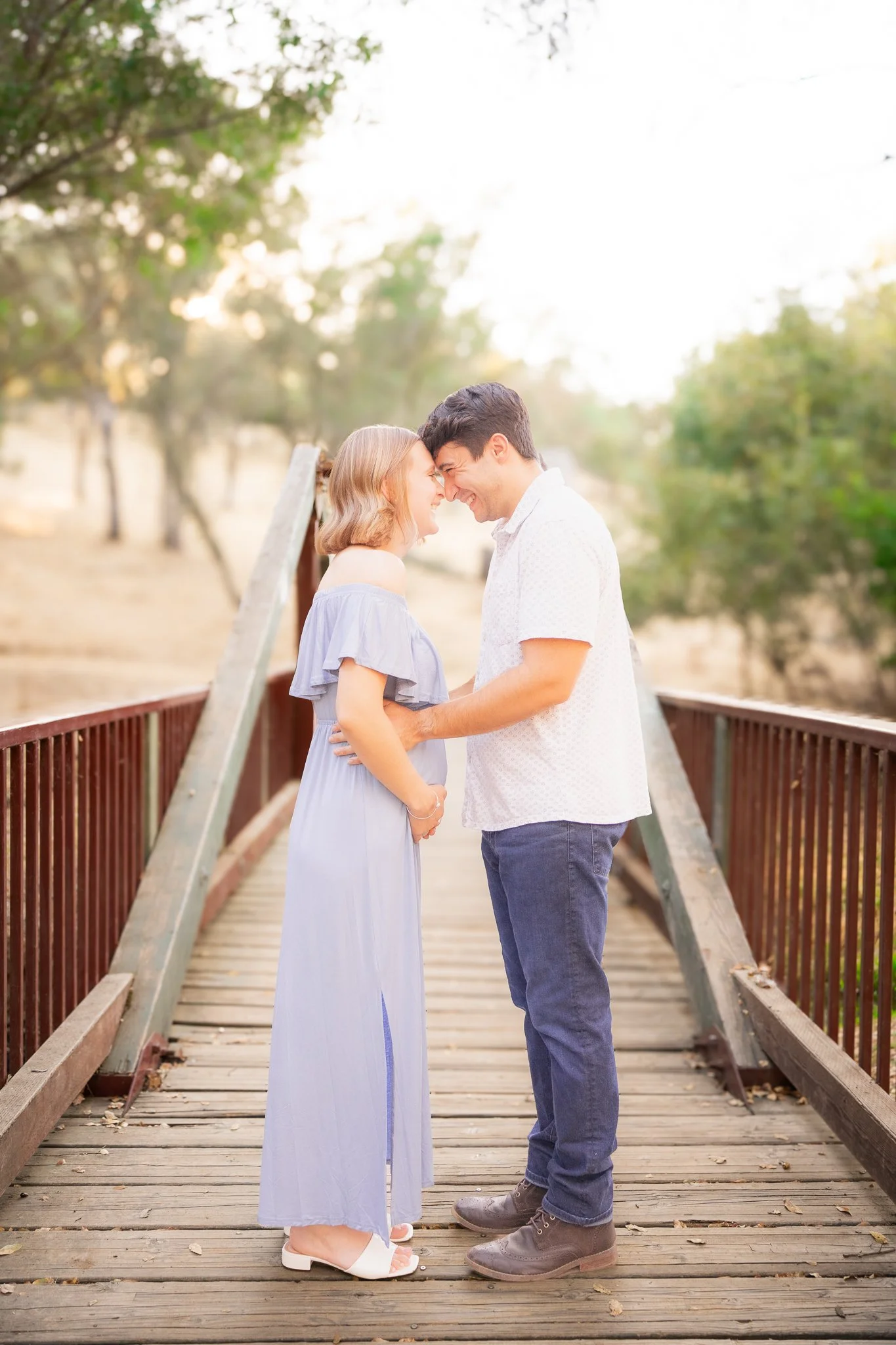 A couple stands face to face on a wooden bridge, smiling and touching foreheads in a park with trees and soft sunlight.