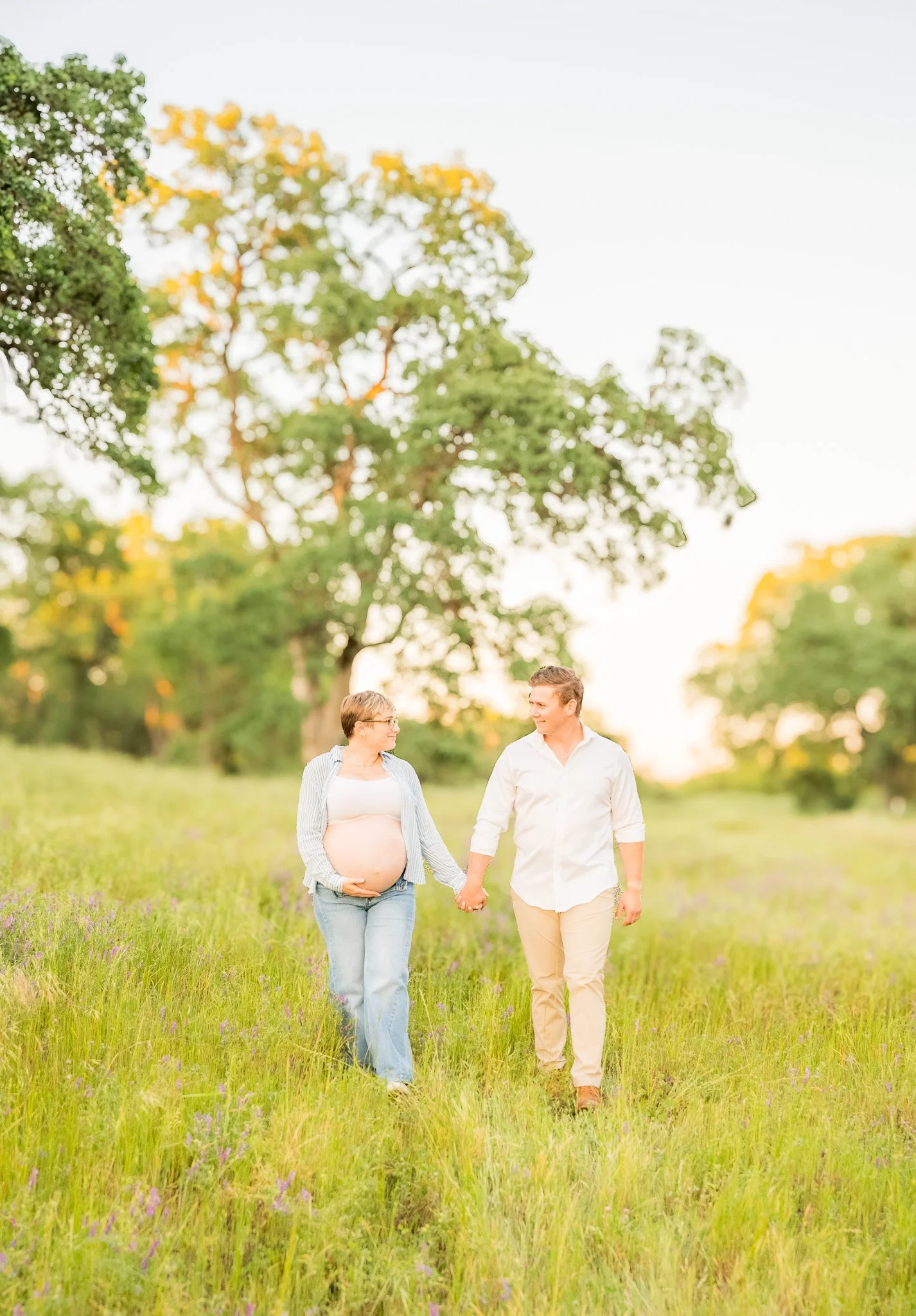 A pregnant woman and a man walking hand in hand through a grassy field with wildflowers, trees in the background, during sunset.