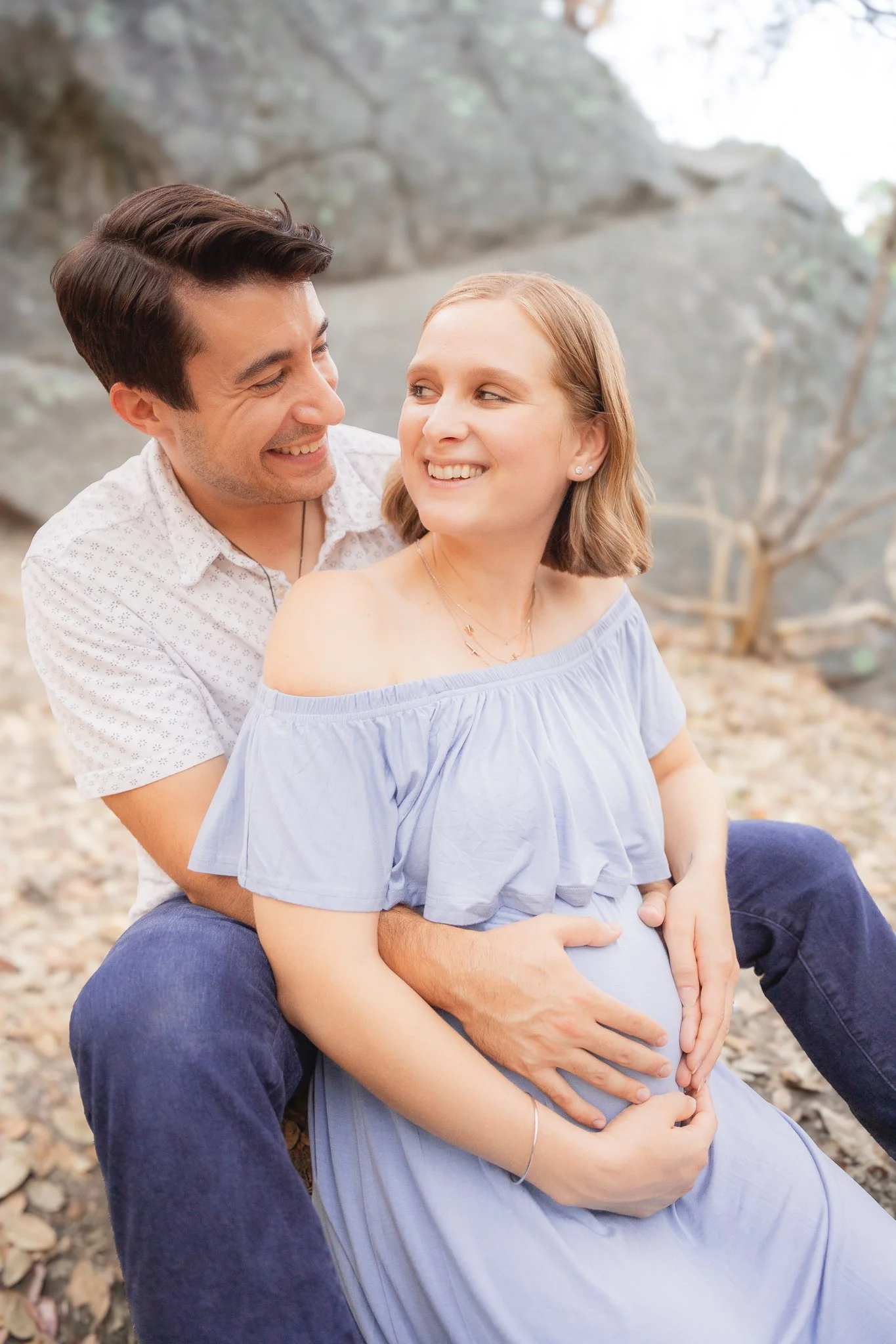 A pregnant woman in a light blue off-shoulder dress sitting outside with a man behind her, both smiling and looking at each other. The man is holding the woman’s pregnant belly with his hands. They are in a natural setting with rocks and trees in the