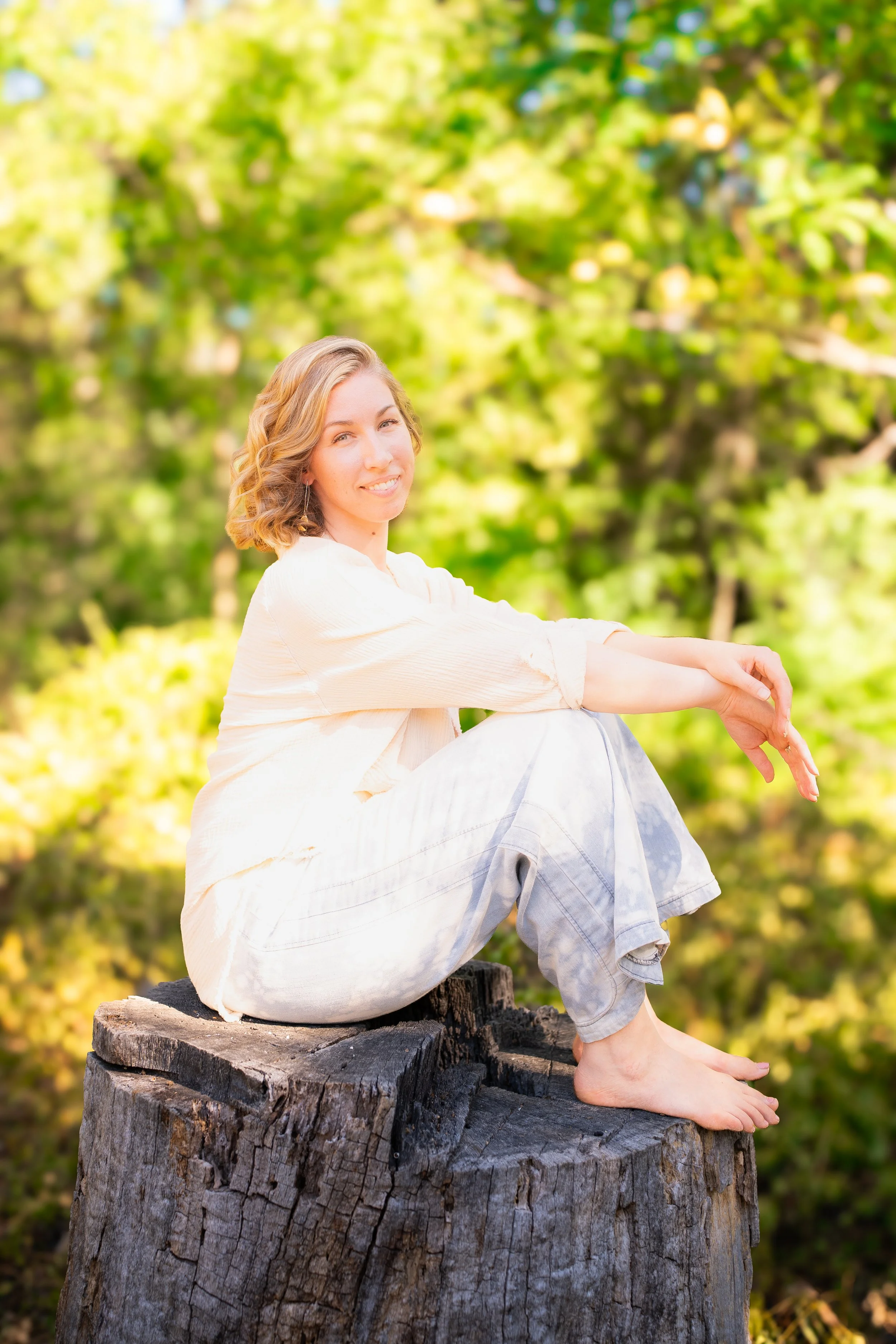 A young woman with wavy blonde hair wearing a light-colored long sleeve top and jeans, sitting barefoot on a tree stump outdoors with green trees in the background, smiling and looking at the camera.