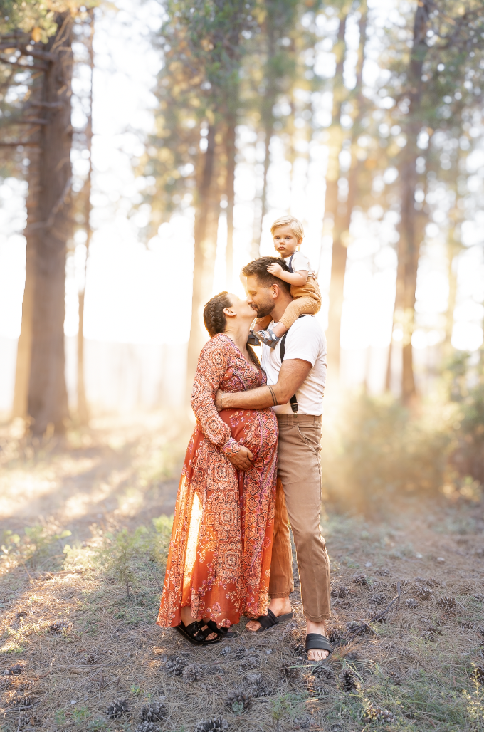 A family of three, including a pregnant woman, a man, and a young child, standing in a forest during sunset. The woman and man are holding hands and smiling at each other while the child sits on the man's shoulders, looking at the camera.