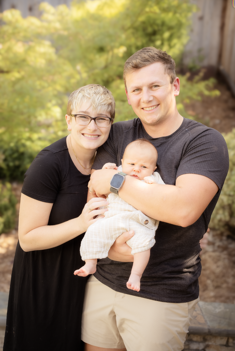 Family of three outdoors, smiling, with a woman with short blonde hair and glasses, a man with short brown hair, and a baby in the man's arms. The background features blurred green and yellow foliage.