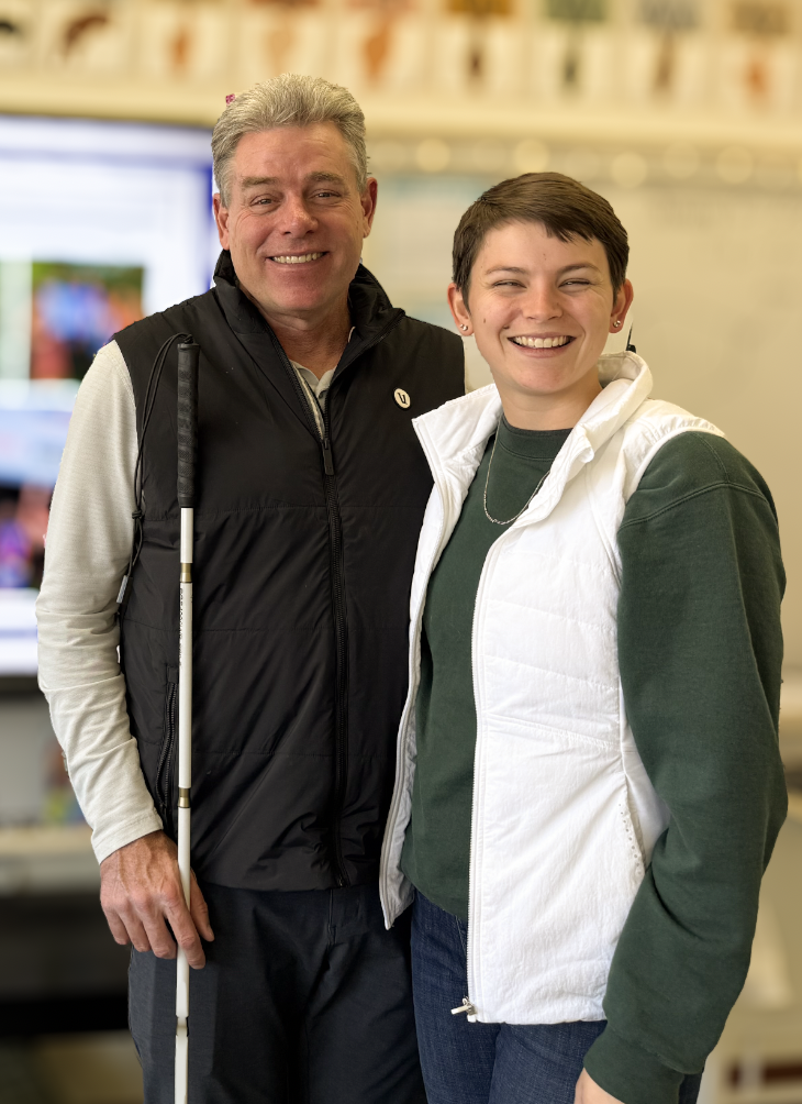 A smiling older man with gray hair, wearing a black vest and holding a golf club, standing next to a young woman with short dark hair, wearing a white vest over a green sweatshirt. They are indoors with a blurred background.
