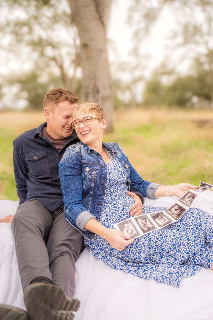 A couple sitting outside, sharing a joyful moment while looking at ultrasound images, with a large tree in the background.