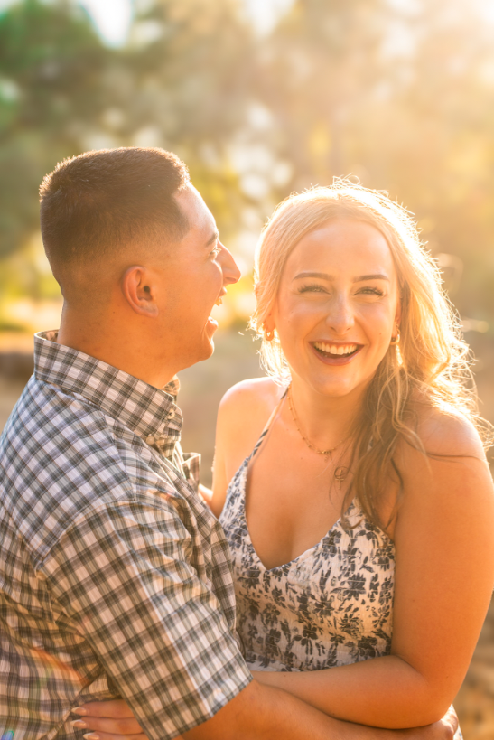 Two young adults, a man and a woman, smiling and laughing outdoors during sunset.