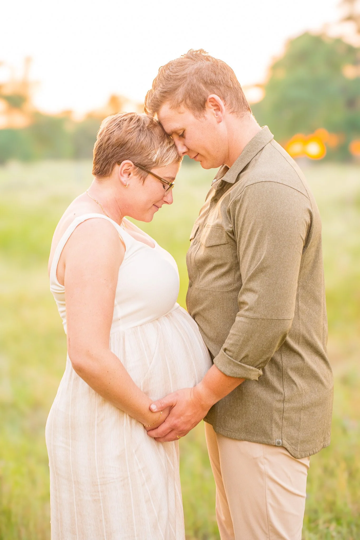 A pregnant woman and a man stand close together with heads touching, holding hands and eyes closed in an outdoor setting during sunset.