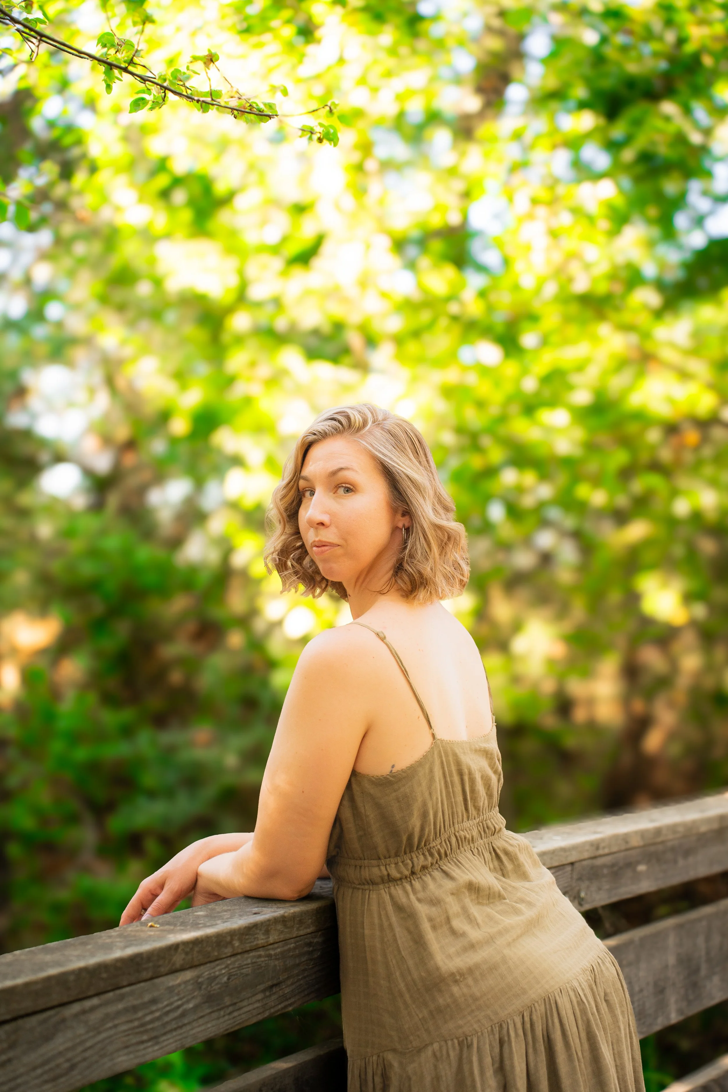 A woman with curly blonde hair wearing a beige dress, sitting on a wooden bench in a park, looking at the camera with a slight smile. Green trees and bright sunlight are in the background.
