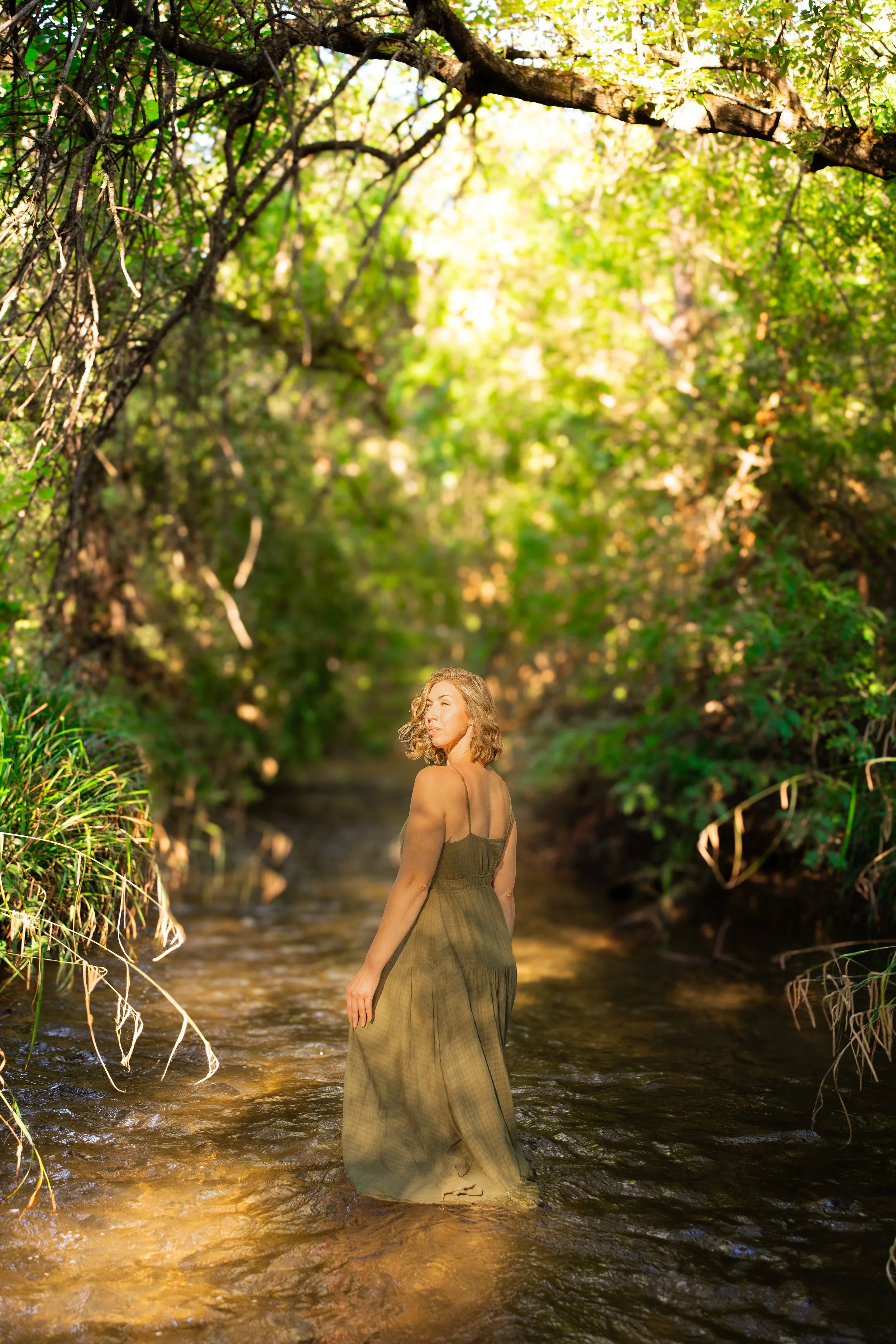 A woman standing in a shallow stream surrounded by green trees and foliage with sunlight filtering through the leaves.
