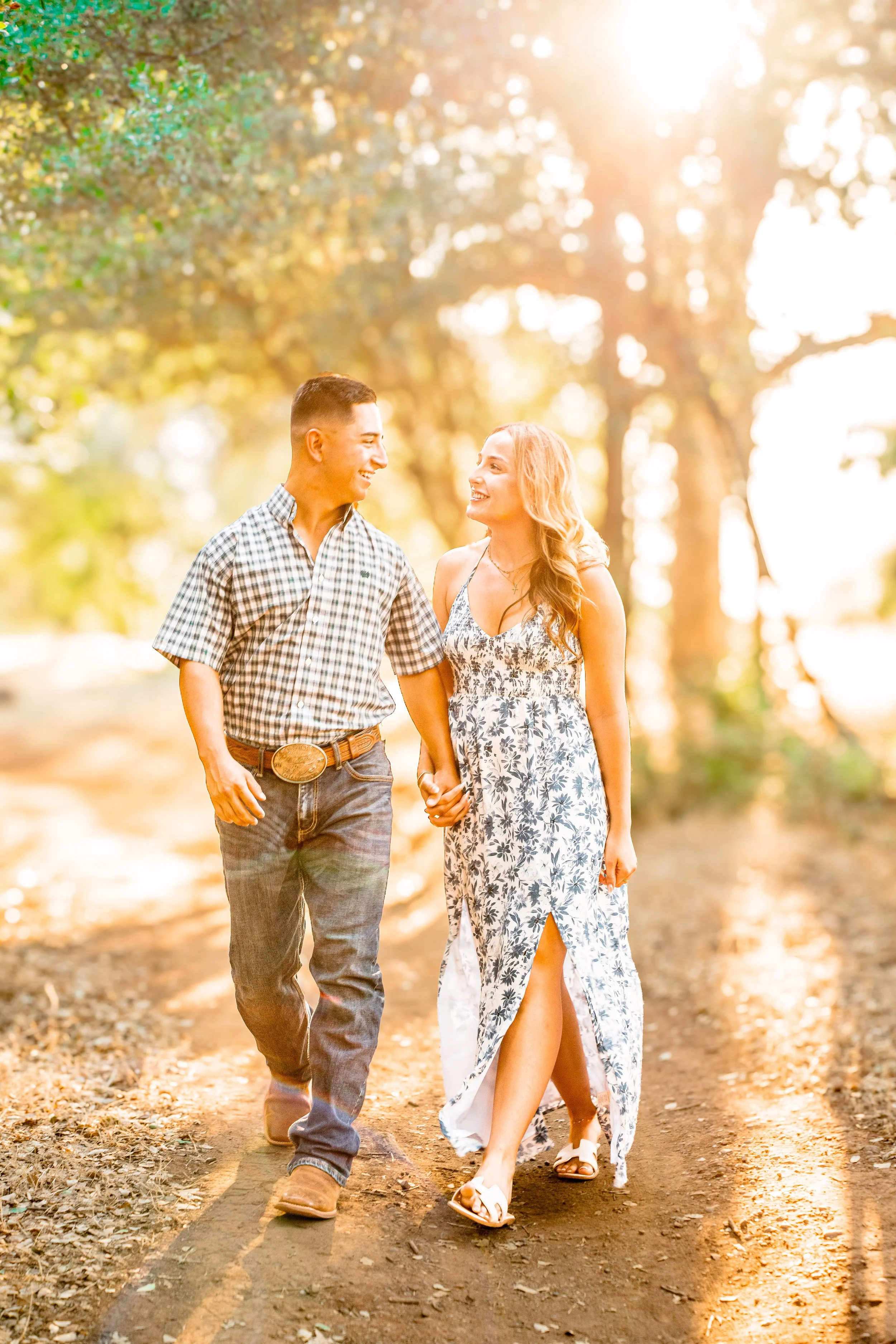 A young couple walking hand in hand on a dirt path in a park during sunset, smiling and looking at each other, with trees and sunlight in the background.