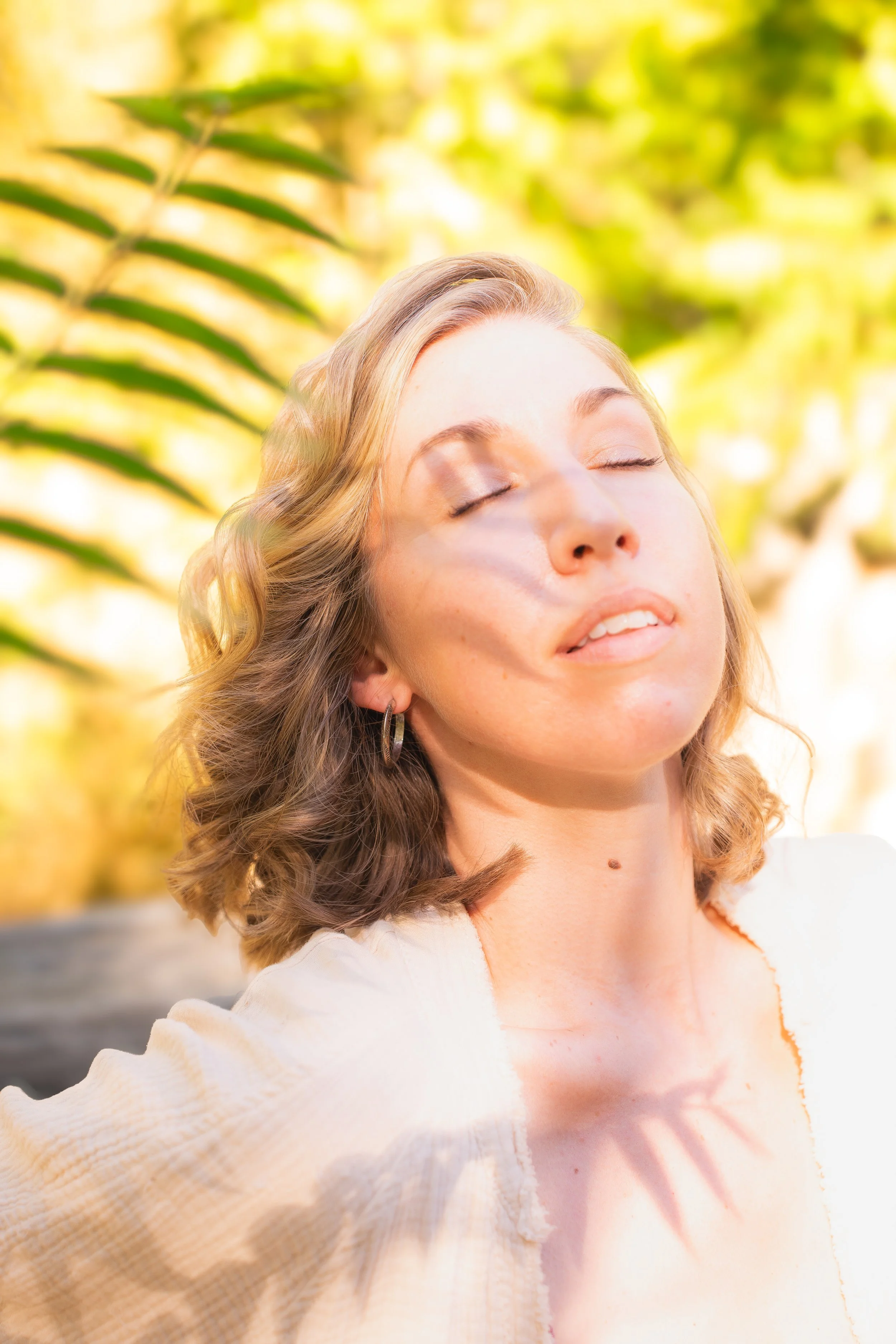 A young woman with wavy blonde hair, closed eyes, and a relaxed expression taking a selfie outdoors on a sunny day. She is wearing a light-colored top, and the background features blurred yellow and green foliage.