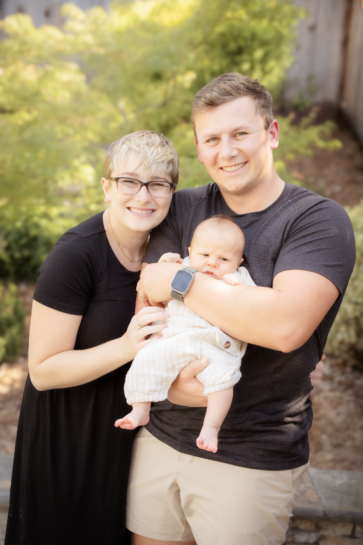 A smiling man and woman holding a baby outdoors with greenery in the background.