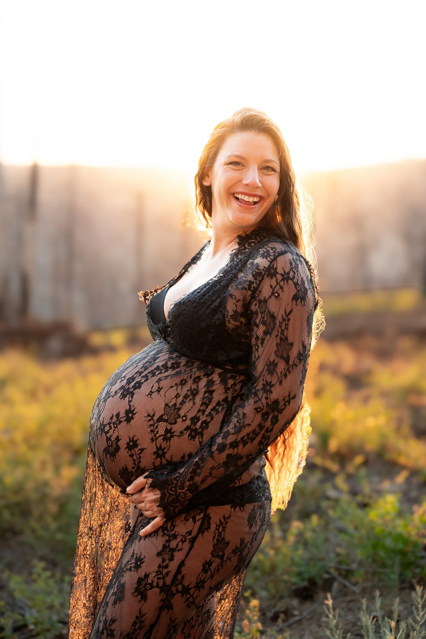A woman with long red hair and a black lace dress standing outdoors during sunset, smiling and holding her pregnant belly.