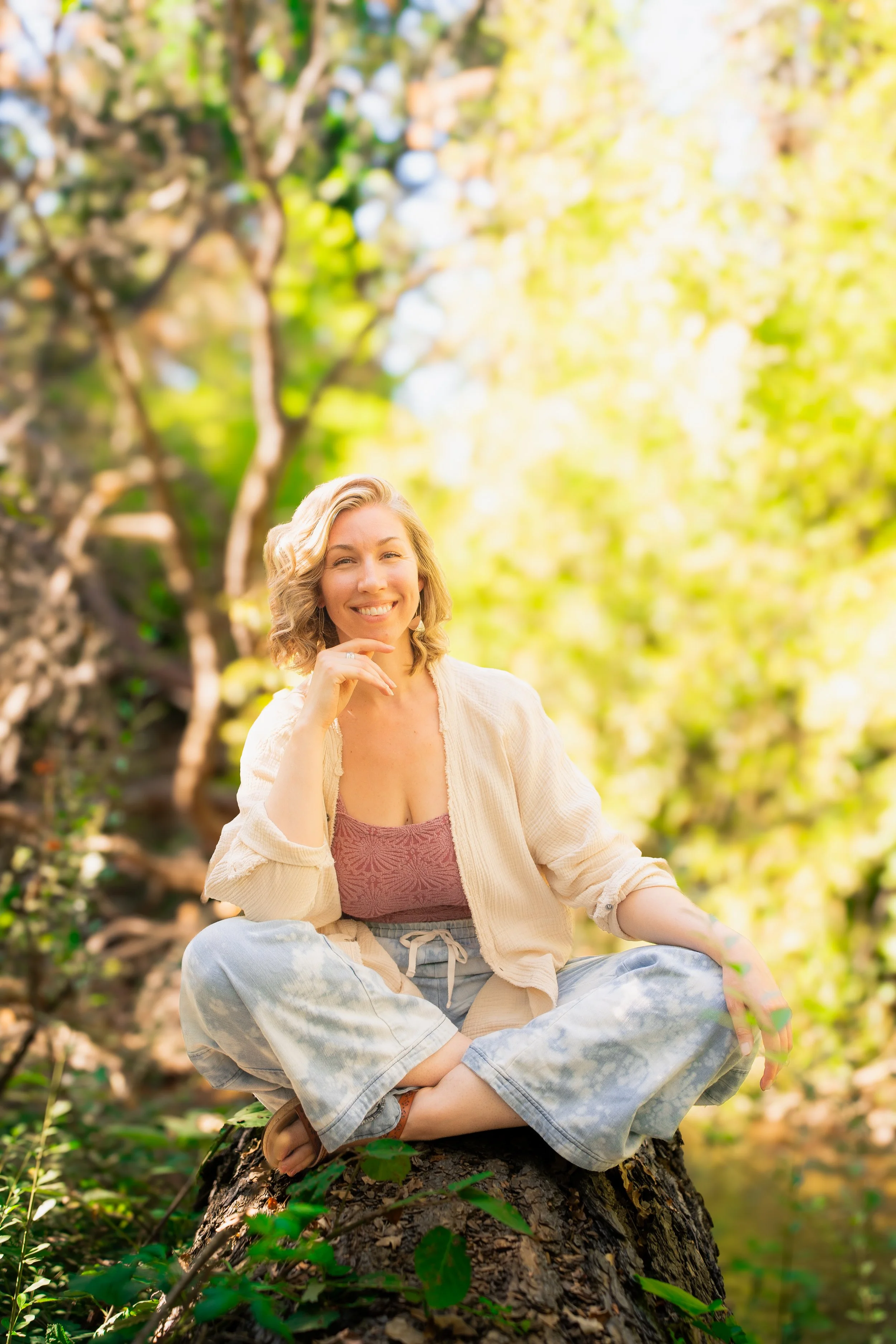 A young woman with blonde curly hair smiling and sitting cross-legged on a tree stump in a green forest during daytime.