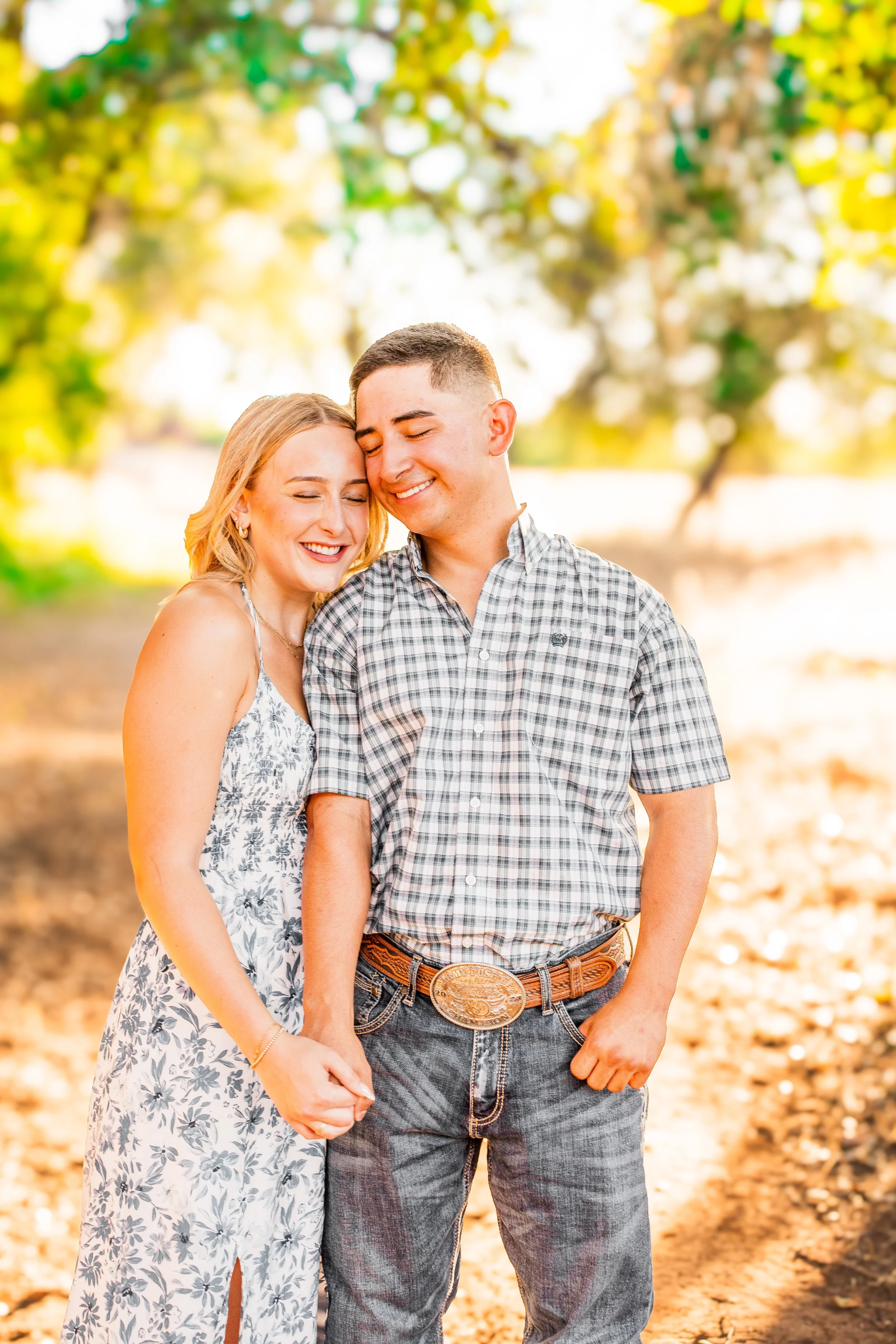 A smiling couple, woman with blonde hair and man with short dark hair, hugging outdoors in sunlight with trees and fallen leaves in the background.