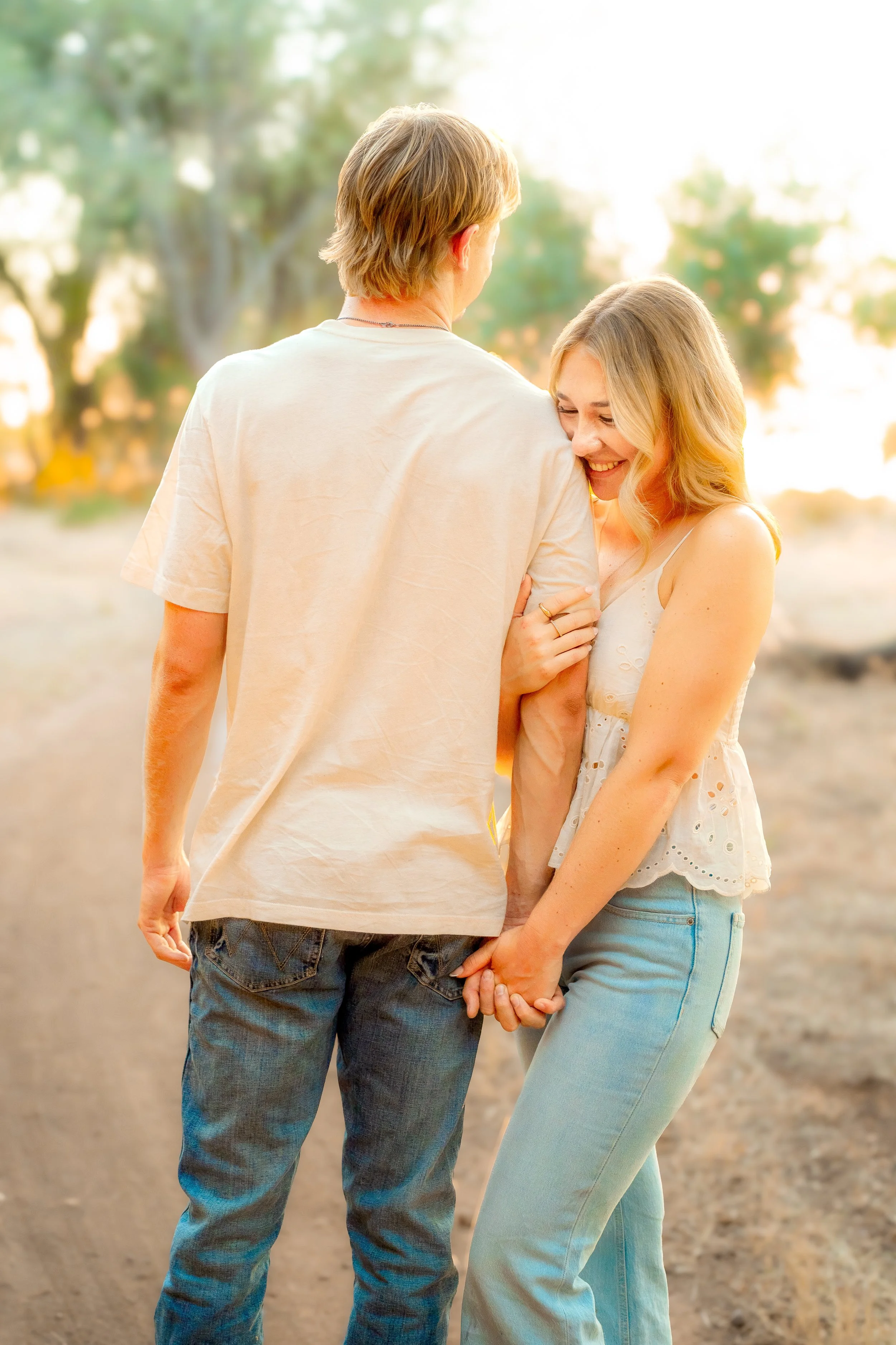 A couple standing close together on a dirt path outdoors, holding hands and smiling, with greenery and trees in the blurred background during a couples session in Bidwell Park in Chico, CA.