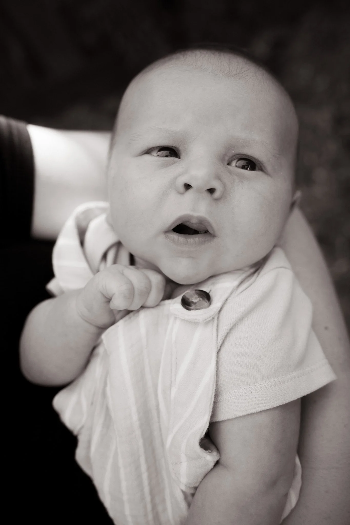 Close-up black and white photo of a baby's face, showing wide eyes, open mouth, and chubby cheeks, with the baby looking directly at the camera.