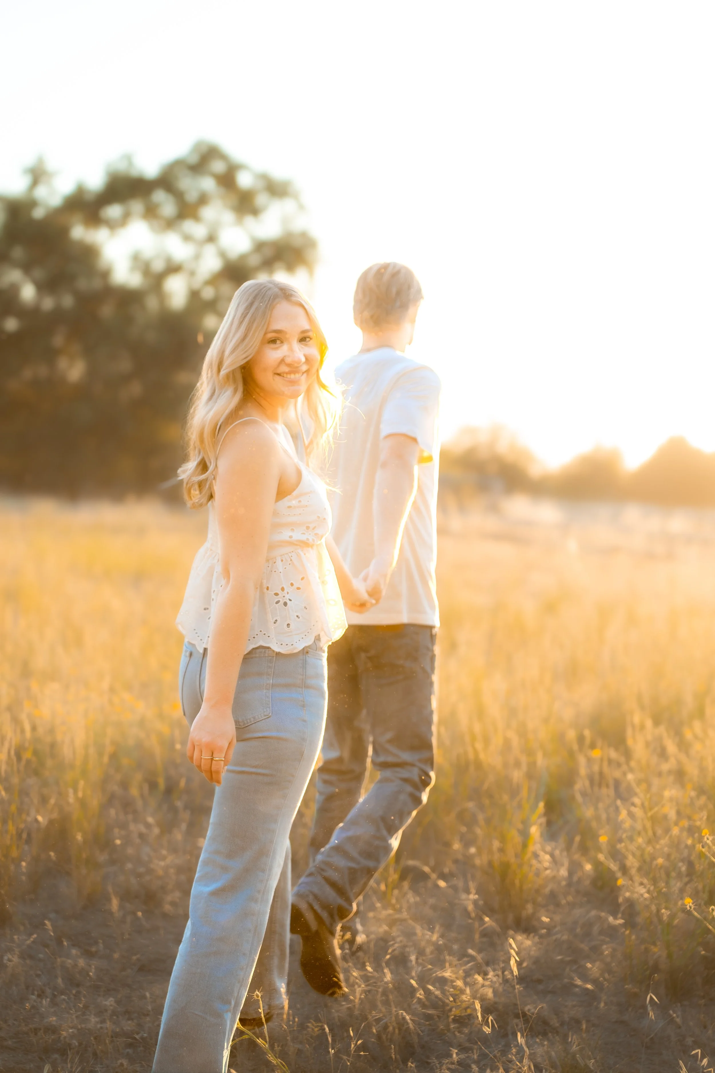 A young woman with blonde hair smiling at the camera holding hands with a man walking away in a field during sunset during a couples session in Bidwell Park in Chico, CA.