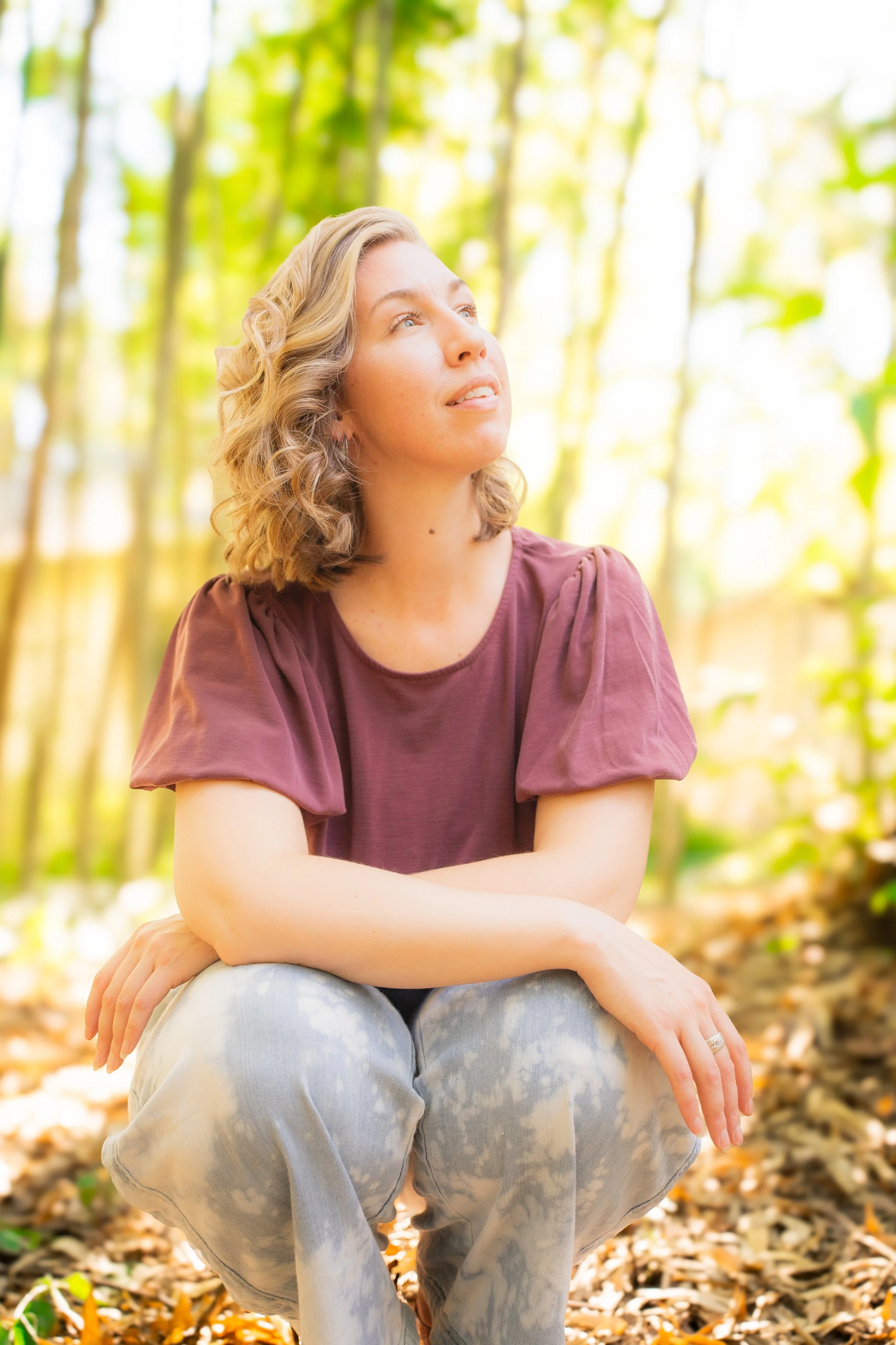 A woman with blonde curly hair sitting outdoors among trees and leaves, looking upward with a serene expression, wearing a purple top and light-colored jeans.