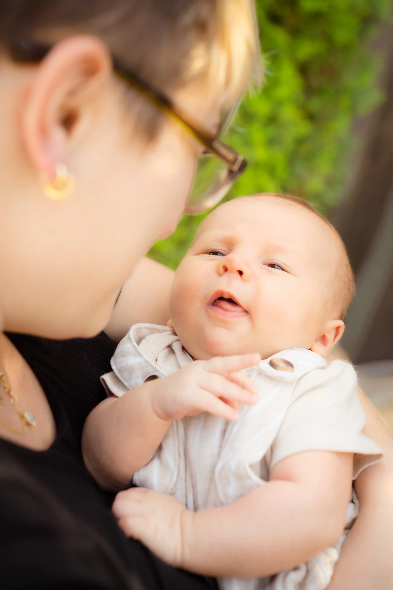A woman wearing glasses and earrings holding a baby in her arms. The baby has a light complexion and is looking up with an open mouth, wearing a cream-colored outfit. The background is green with blurred foliage.