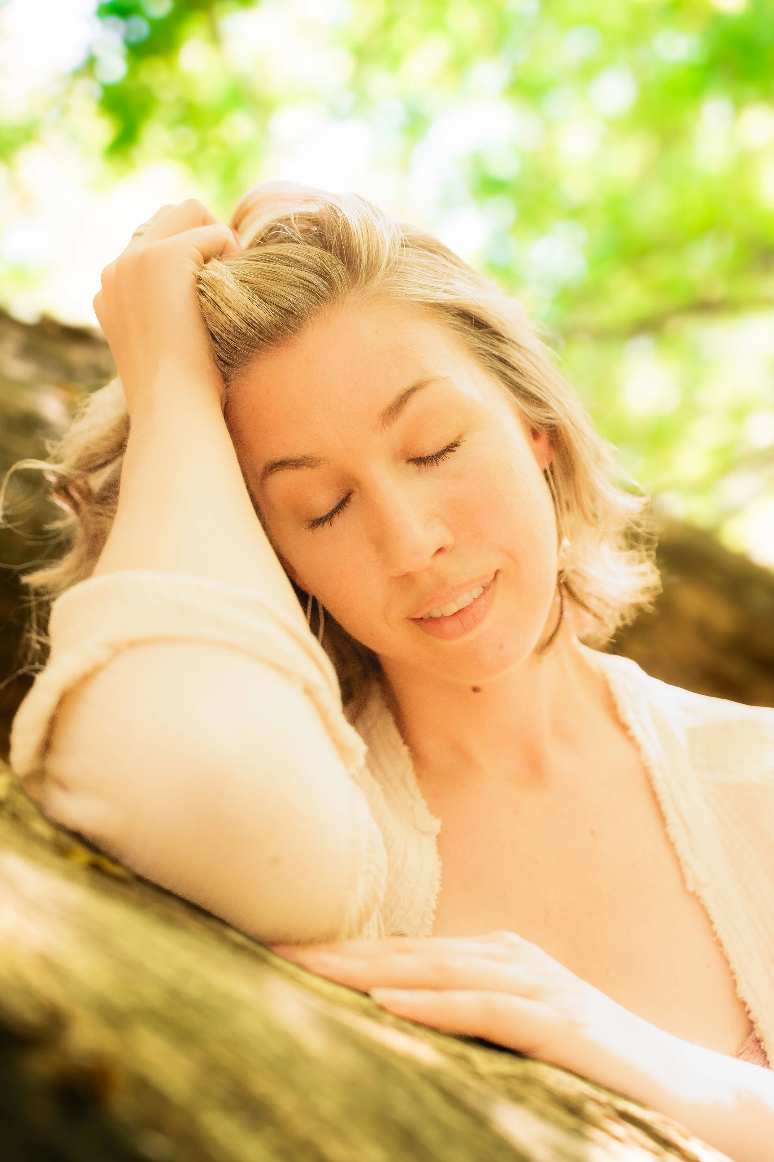 A woman with closed eyes, smiling gently, resting her head and hand on a tree branch with blurred green foliage in the background.