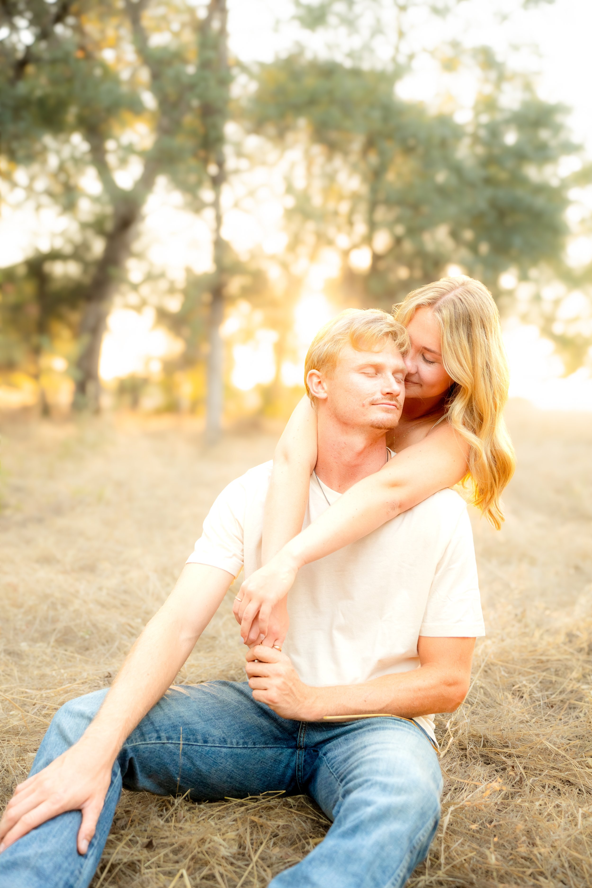 A couple sitting outdoors on dry grass, with the woman hugging the man from behind during a couples session in Bidwell Park in Chico, CA.