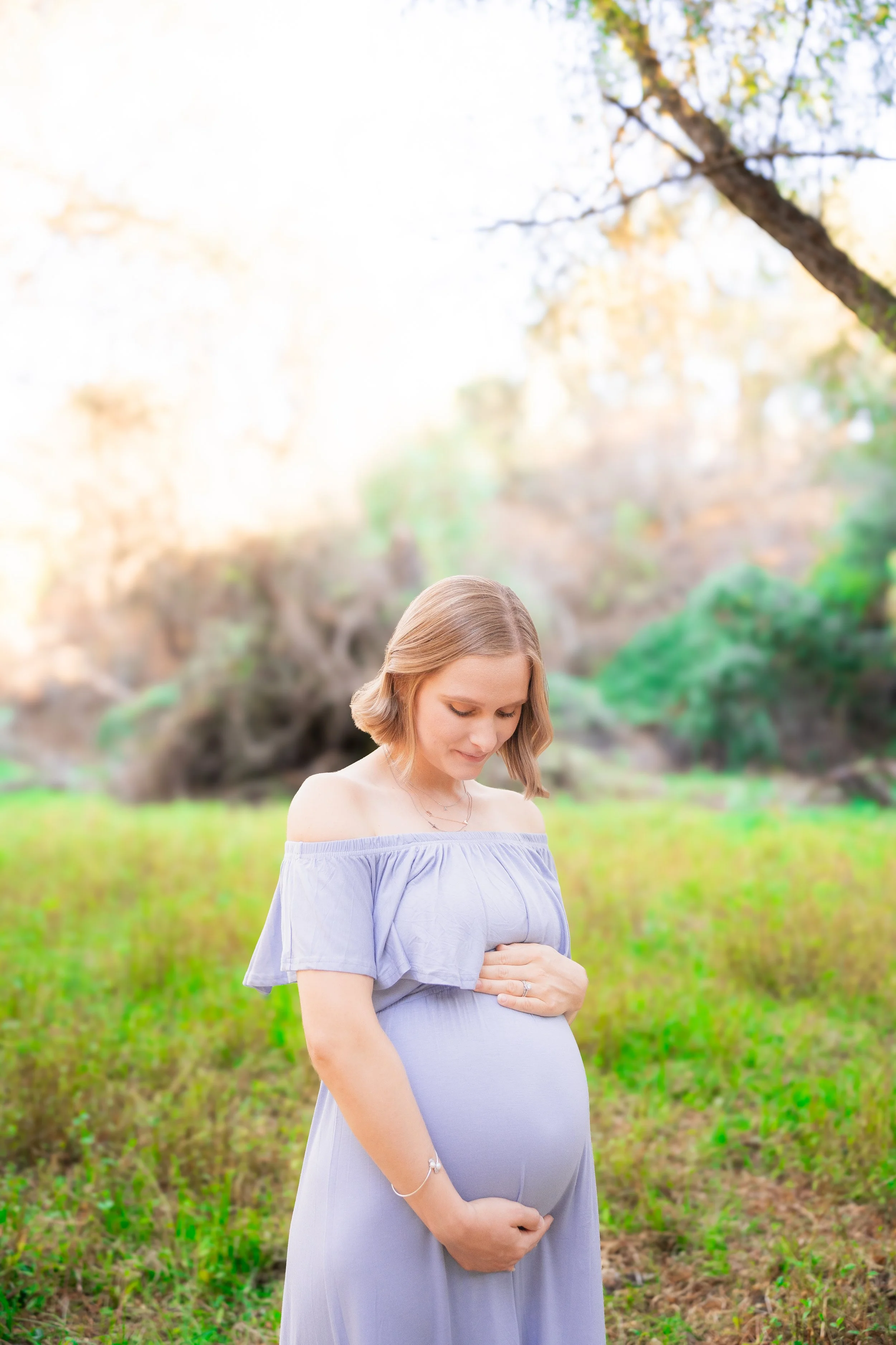 A pregnant woman with light brown hair wearing a lavender off-shoulder dress, gently holding her baby bump with both hands, standing outdoors in a park with green grass and trees in the background.