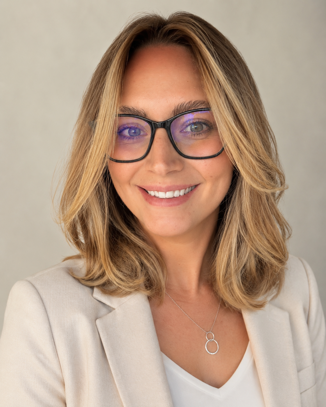 A woman with shoulder-length blonde hair, wearing glasses, a beige blazer, and a white top, smiling at the camera against a neutral background.